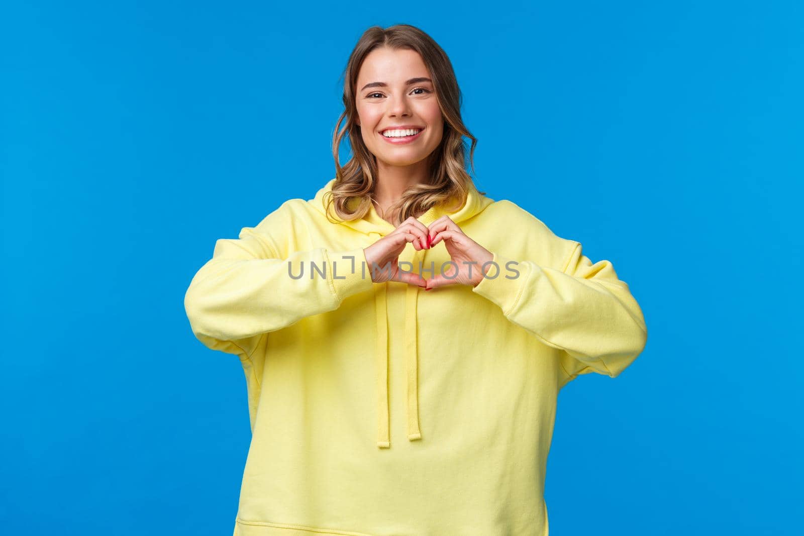 Lovely caring cute blonde girl in yellow hoodie showing heart gesture and smiling as express symapthy, confess in love or heartwarming feelings towards person, stand blue background by Benzoix
