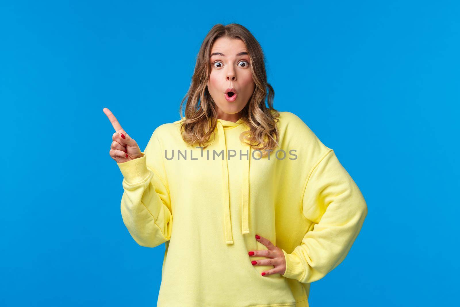 Wow incredible. Impressed and fascinated blond caucasian female pointing finger upper left corner and look camera astonished, telling about awesome event, standing blue background.