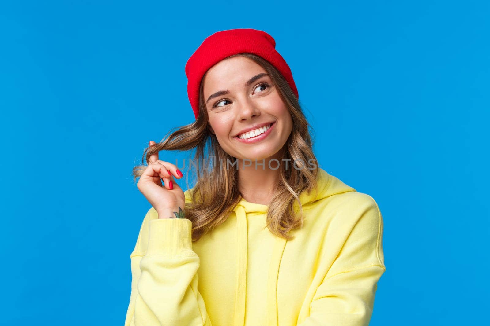 Close-up portrait dreamy silly cute european female in red hipster beanie, yellow hoodie, rolling curl on hair and gazing upper right corner thoughtful, standing blue background. Copy space