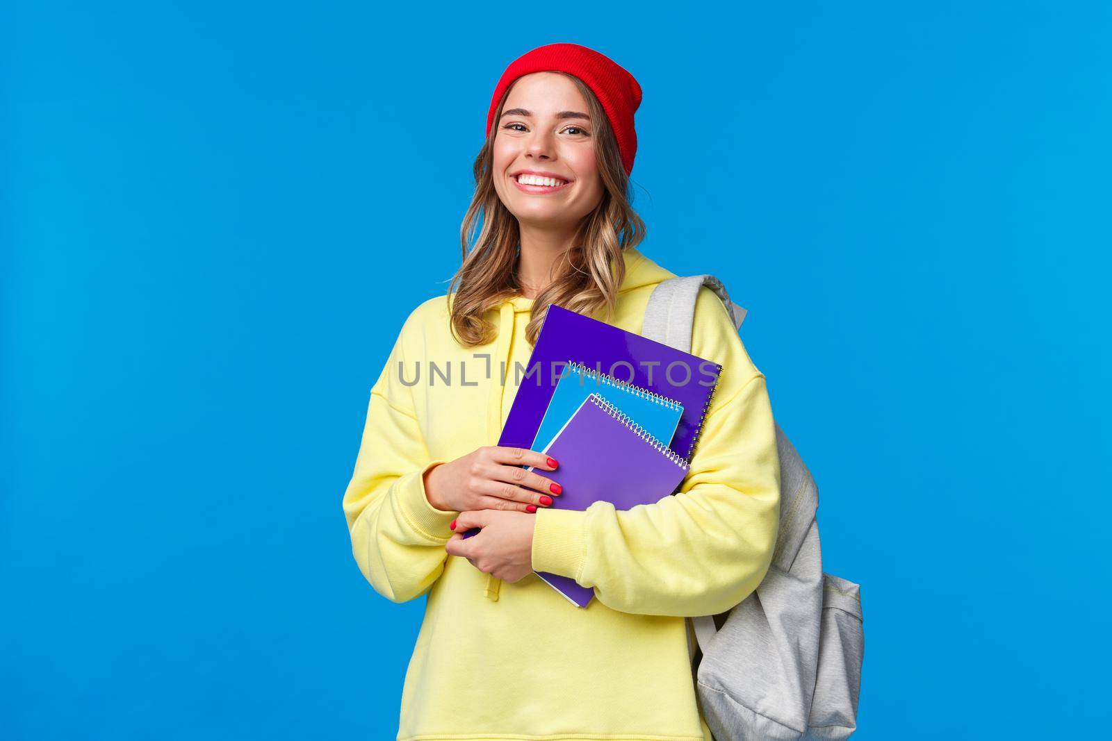 Charismatic smiling female in red beanie and yellow hoodie, walking to college, carry backpack and notebooks, papers for classes, look joyfully, studying prepare for exams to enter university.