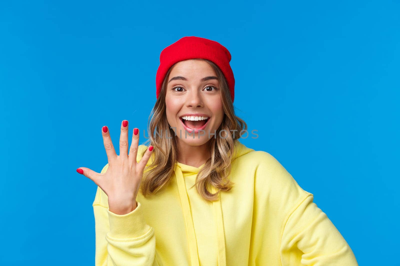 Close-up portrait of good-looking cheerful smiling woman in red beanie and yellow hoodie, showing number five or fifth, make order, reserve seats for friends, stand blue background.