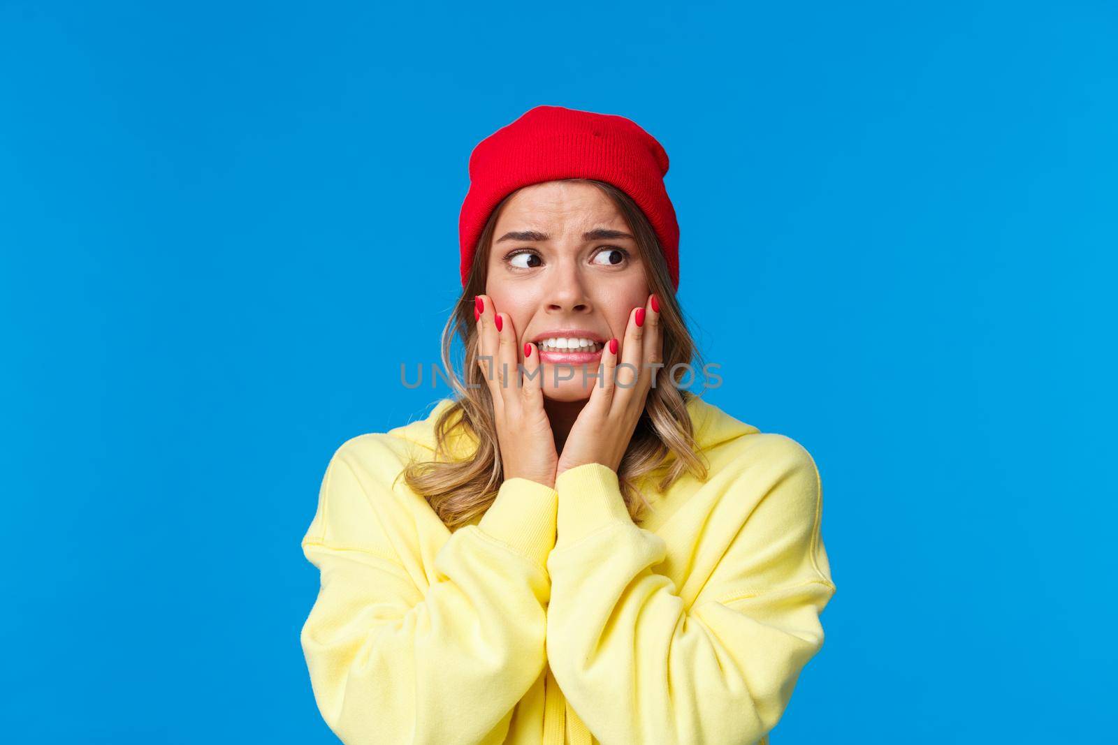 What have I done. Worried and concerned anxious caucasian female in red beanie and yellow hoodie, hold hands on cheeks panicking look away frightened, standing troubled blue background.