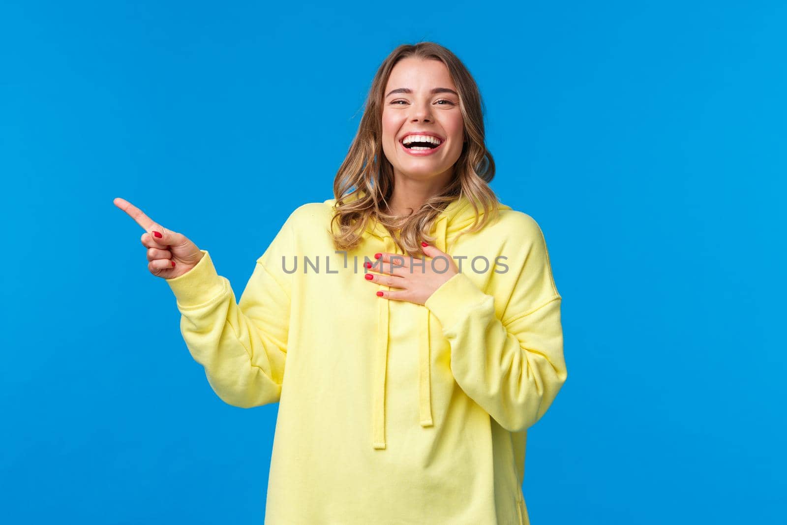 Happy and amused good-looking blond female in yellow hoodie, laughing out loud over something hilarious on upper left corner of blue background, smiling and looking entertained camera.