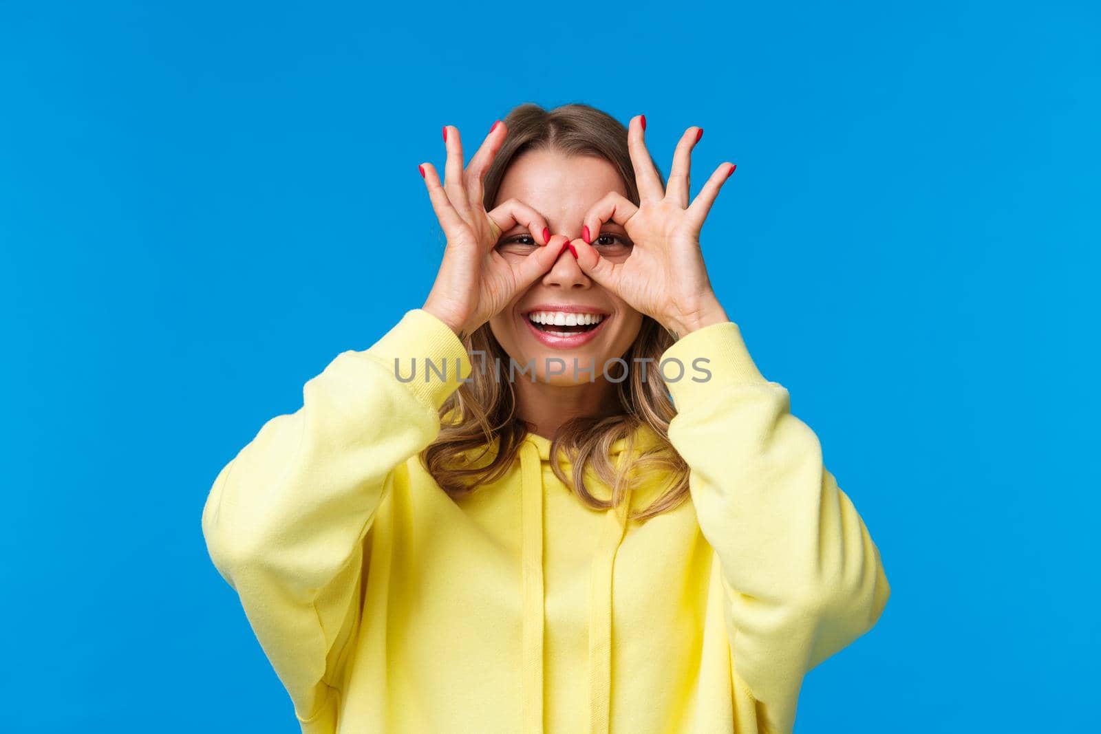 Close-up portrait cheerful and playful cute blonde girl making glasses with fingers, looking through it with amused happy and entertained grin, standing carefree over blue background.