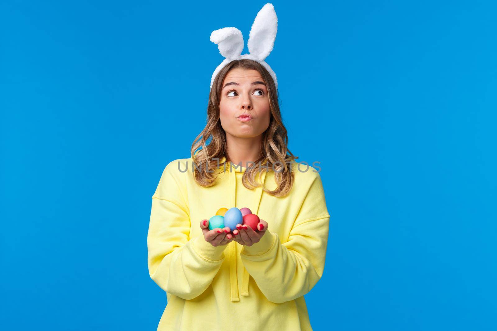 Thoughtful cute caucasian girl in rabbit ears thinking, looking up pondering and dreamy, holding Easter eggs, decide where hide it during traditional holiday game, blue background by Benzoix