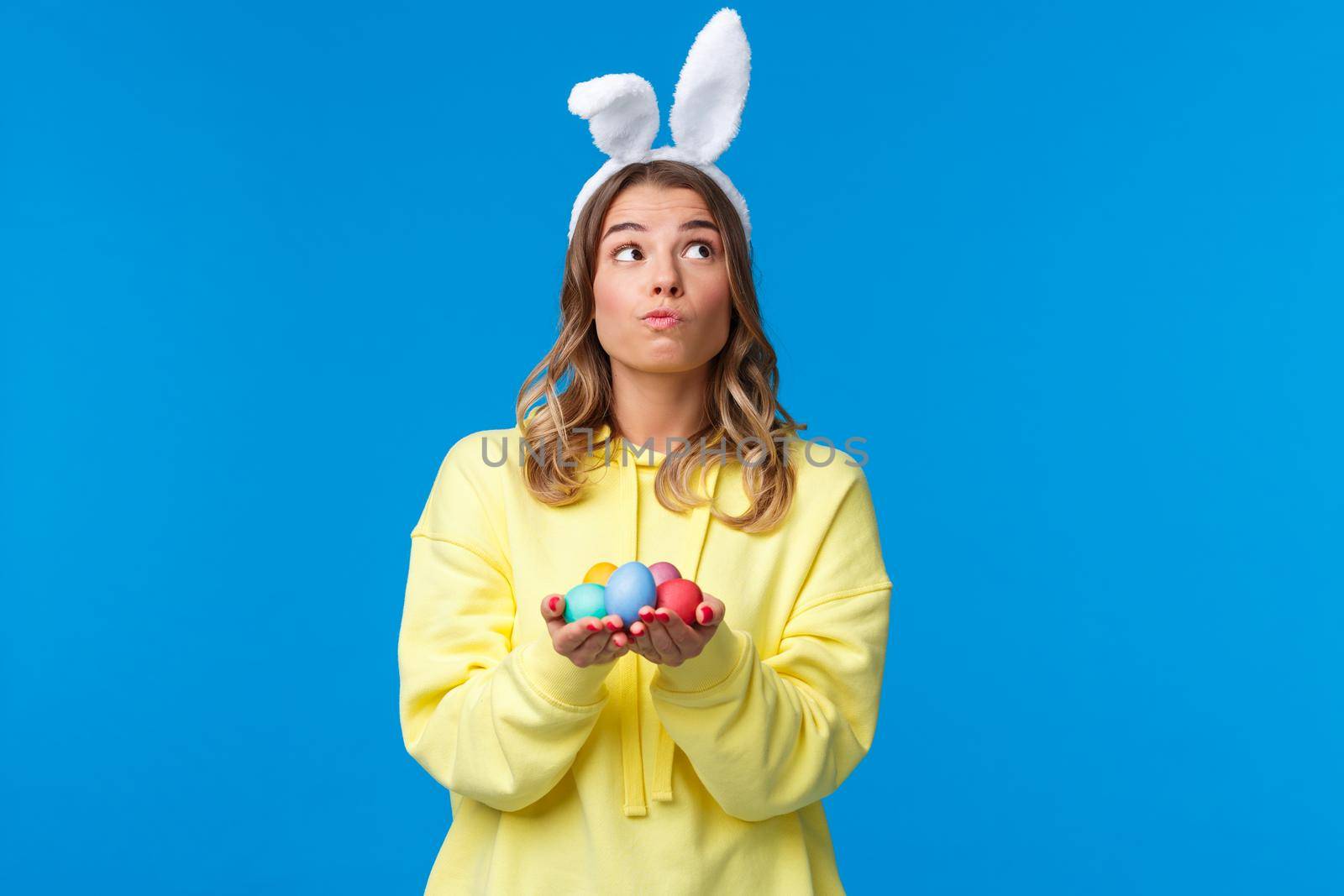 Thoughtful cute caucasian girl in rabbit ears thinking, looking up pondering and dreamy, holding Easter eggs, decide where hide it during traditional holiday game, blue background.