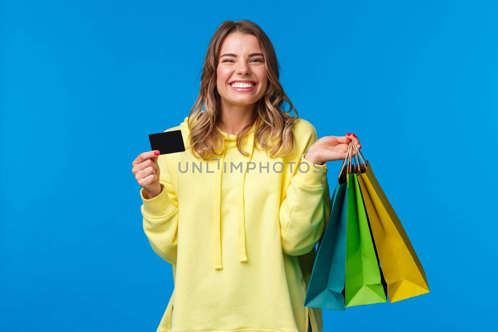 Happy smiling pretty girl using her credit card, deposit money for shopping, holding bags with clothes and grinning delighted, finally getting ready for summer vacation, blue background by Benzoix