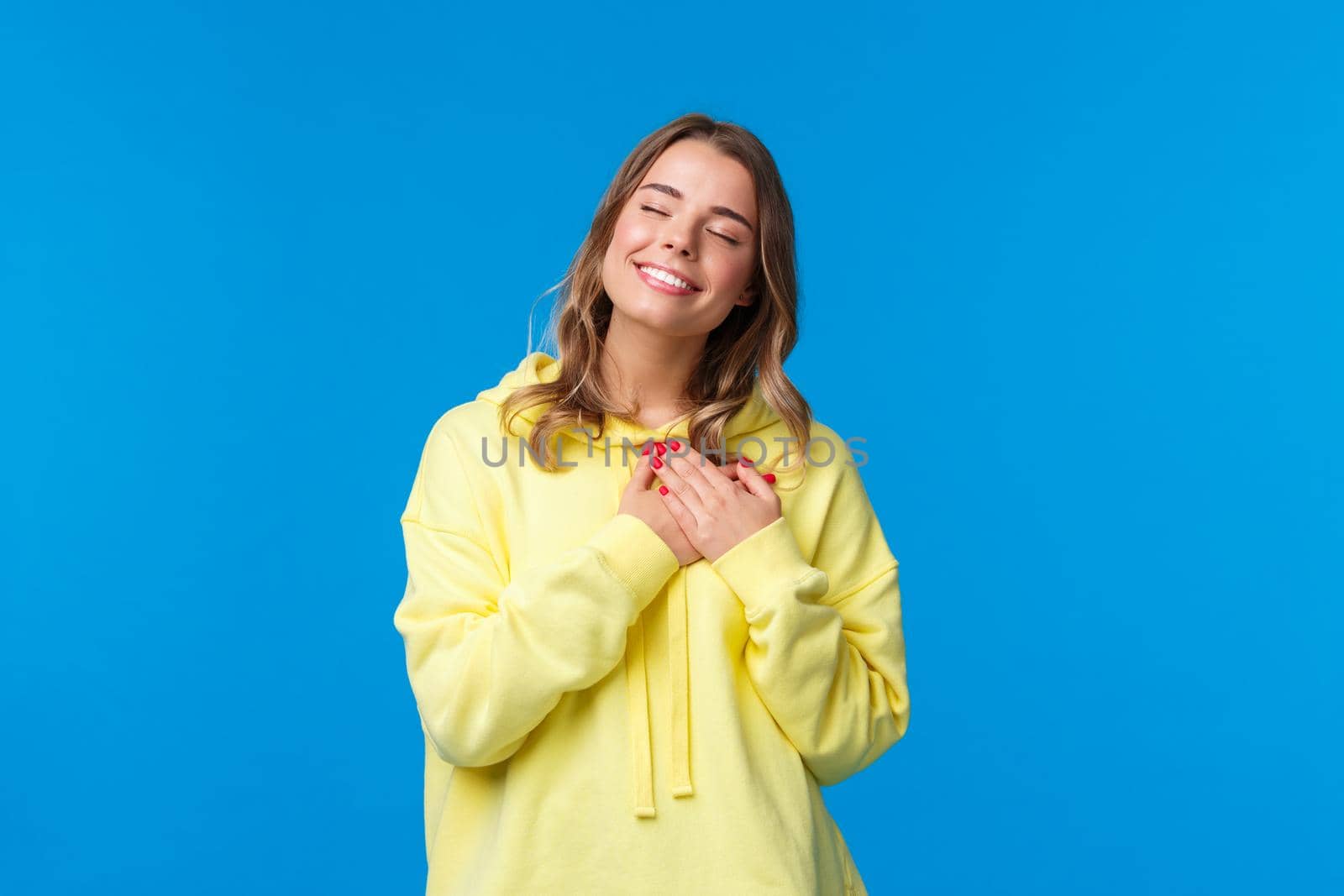 Romantic alluring blond caucasian girlfriend in yellow hoodie, press hands to heart and close eyes with lovely smile recall beautiful memory, imaging perfect date, standing blue background by Benzoix