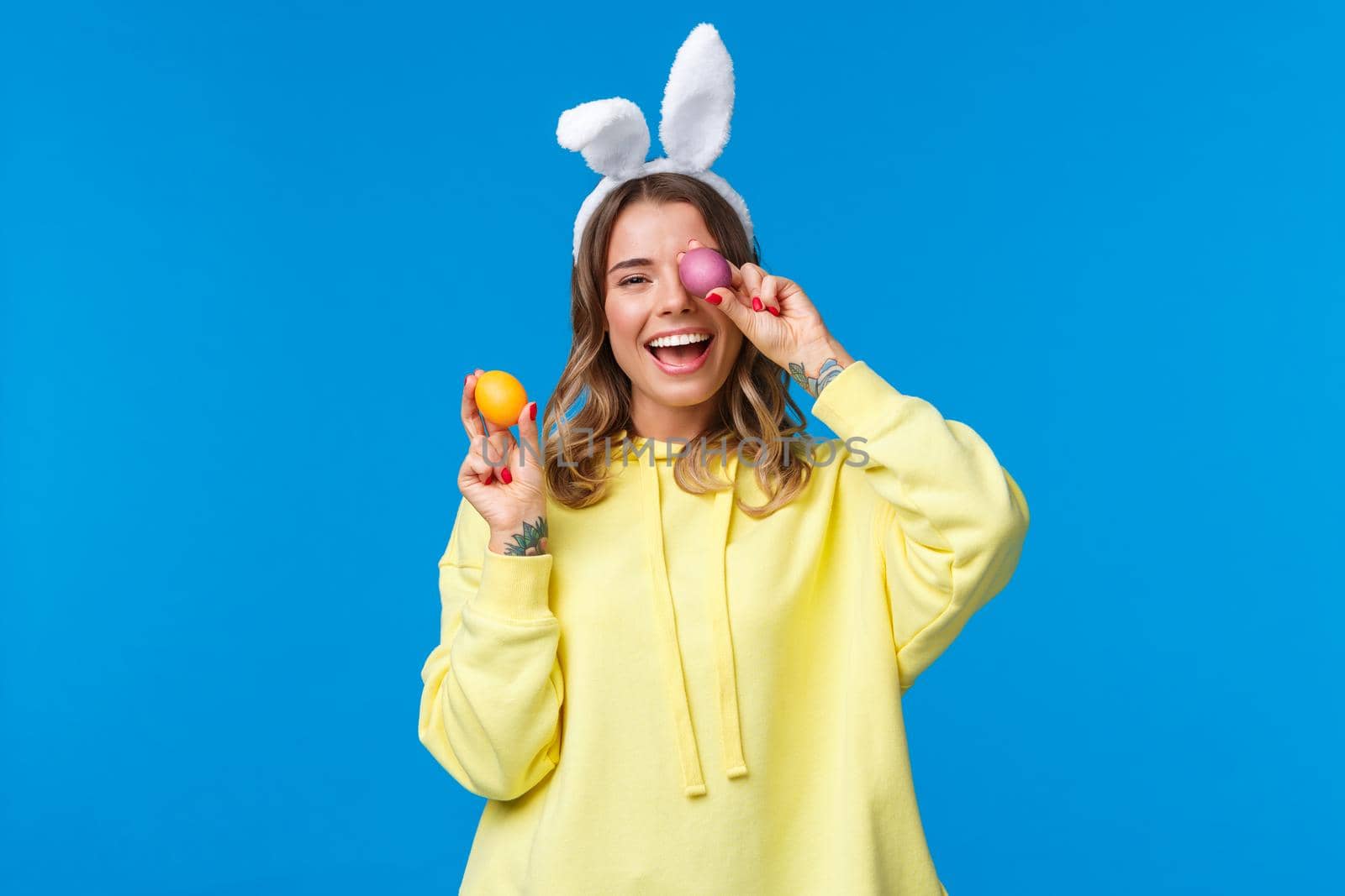 Holidays, traditions and celebration concept. Happy cheerful young pretty female celebrating Easter day, showing two painted eggs and laughing, wearing cute rabbit ears, blue background.