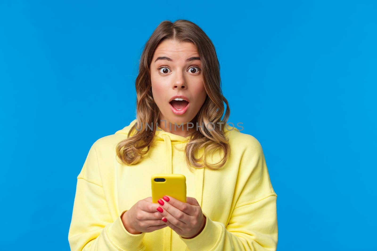 Close-up portrait entertained and surprised cute blond girl look with disbelief and amazement camera after reading interesting news online, holding mobile phone, standing blue background by Benzoix