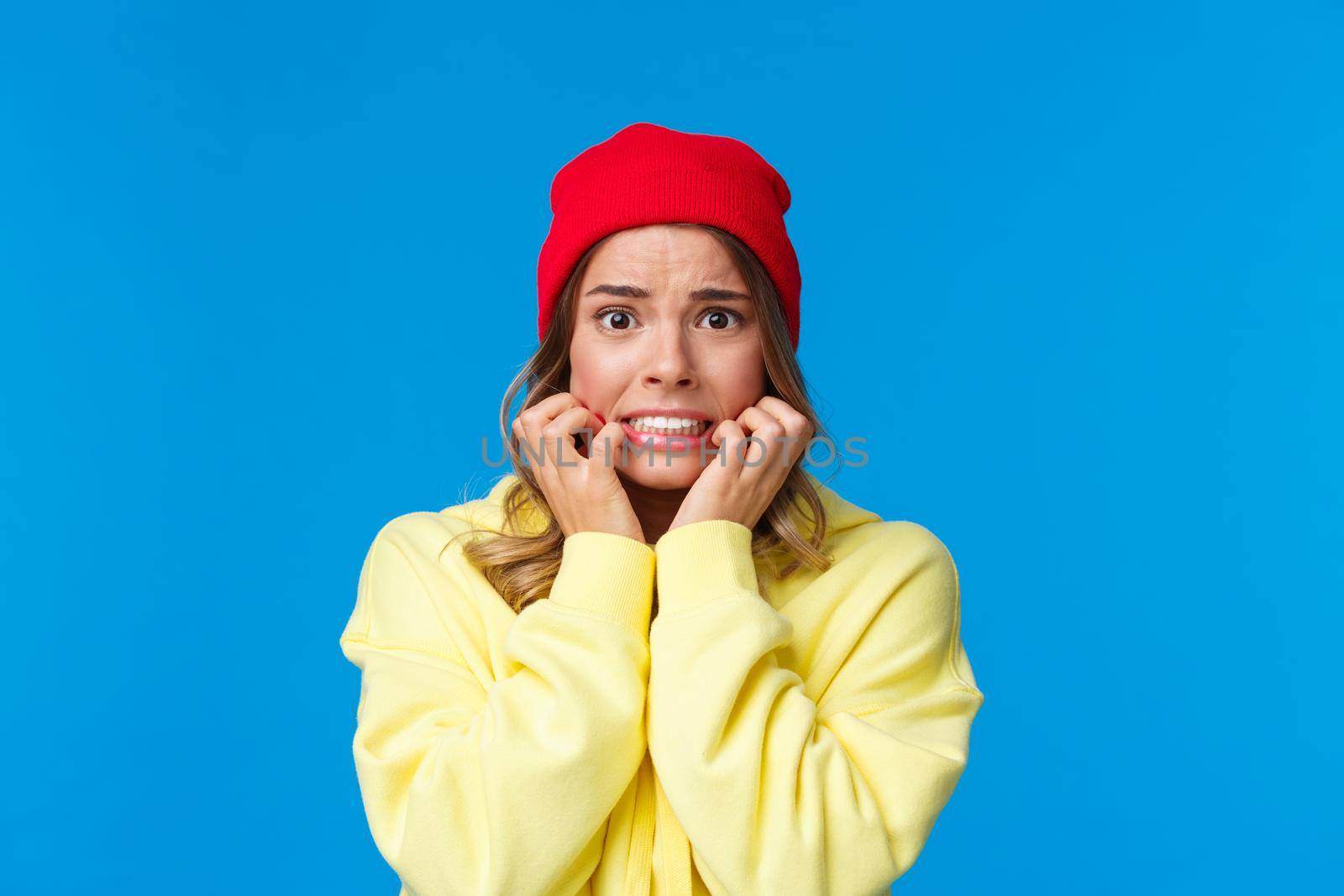 Close-up portrait of scared and insecure cute troubled caucasian girl in red beanie, biting fingernails and frowning concerned, look afraid and worried over some trouble, standing blue background.
