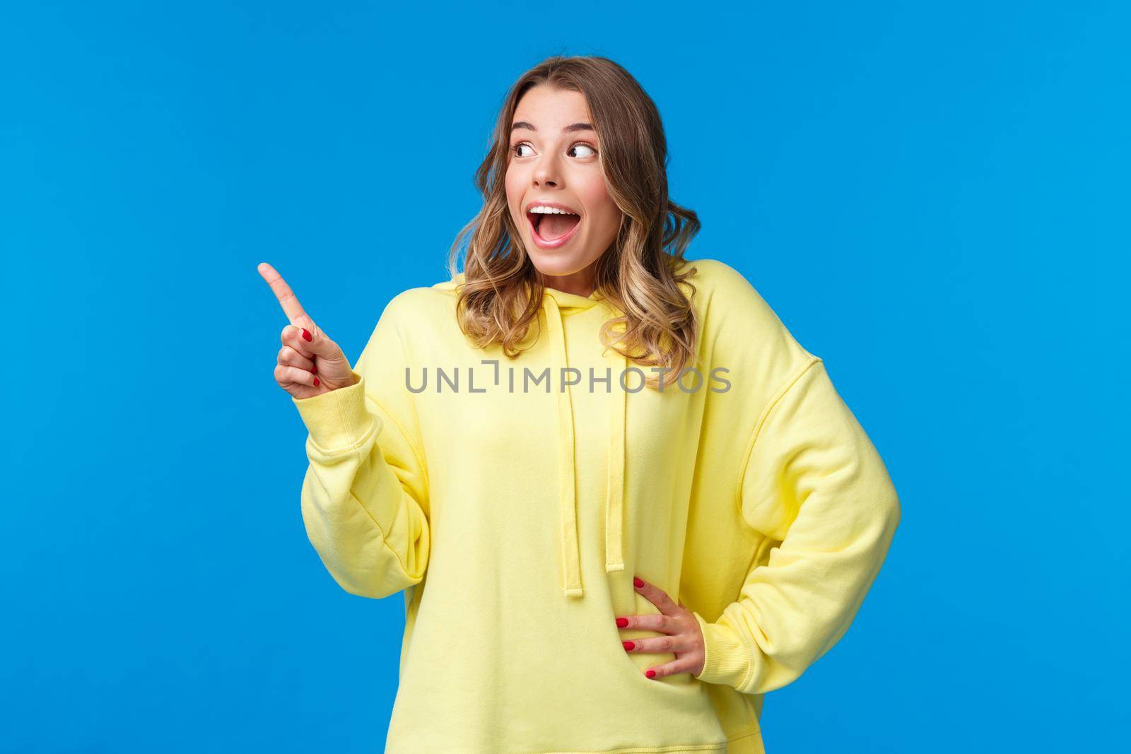 Excited speechless happy blond girl react to awesome news, pointing and staring at left blank copy space as if seeing something stunning and cool, standing blue background by Benzoix