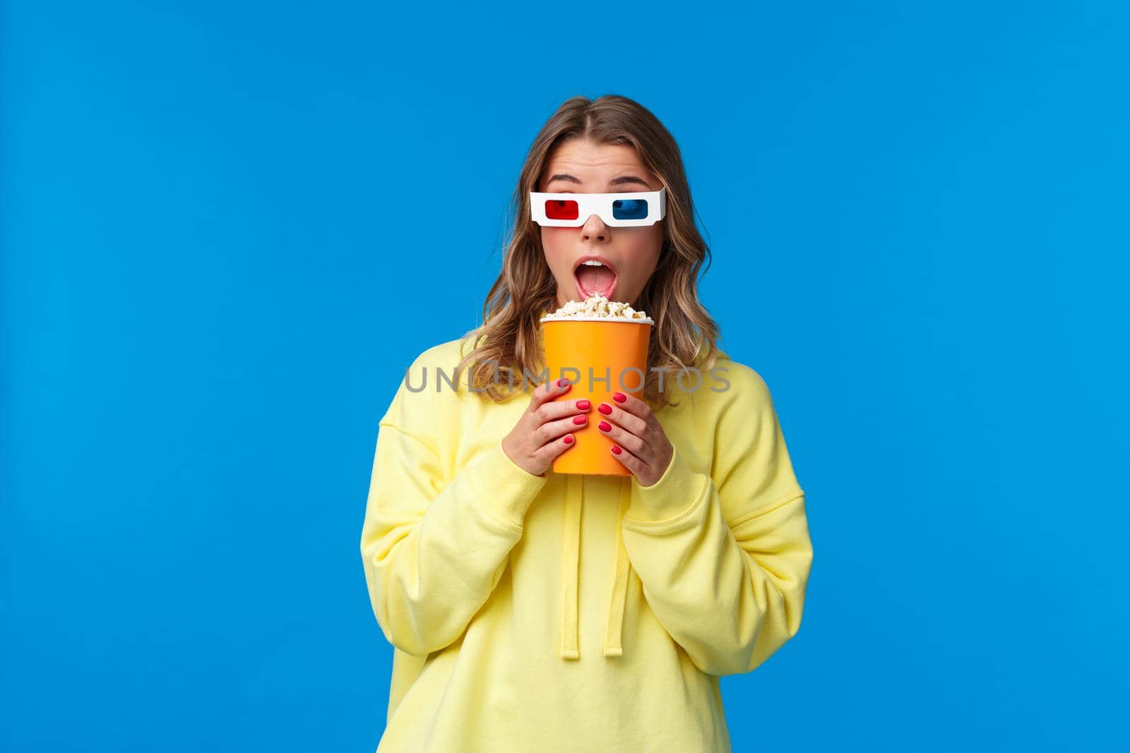 Leisure, fun and youth concept. Cheerful pretty blond young woman in yellow hoodie eating popcorn look away and wear 3d glasses as watching movie in cinema, blue background.