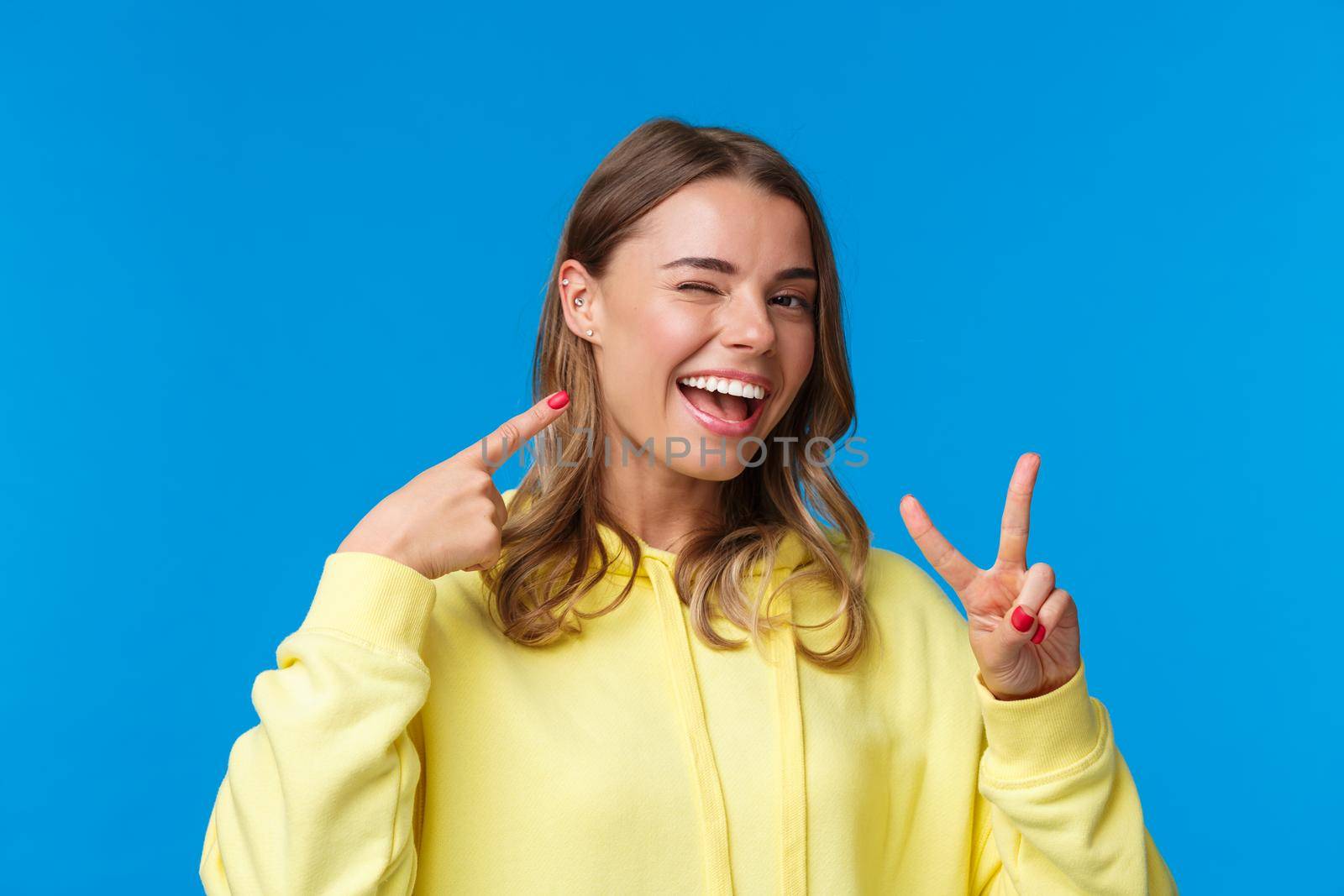 Close-up portrait of proud and boastful cute blond girl with short hair and pierced ear, pointing at herself and make peace gesture with pleased smile, standing blue background upbeat.