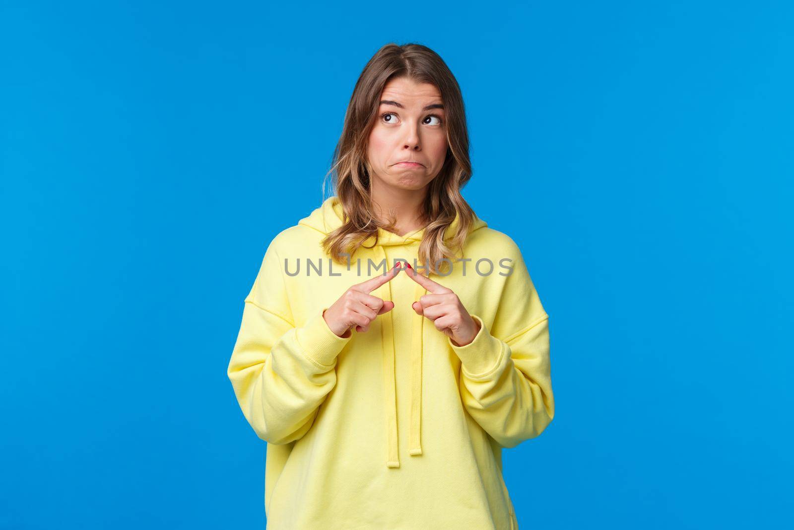 Shy and silly kawaii blond girl in yellow hoodie being modest embarrassed say I love you, poking fingers and look away not to have eye contact as blushing, have something to ask, blue background.