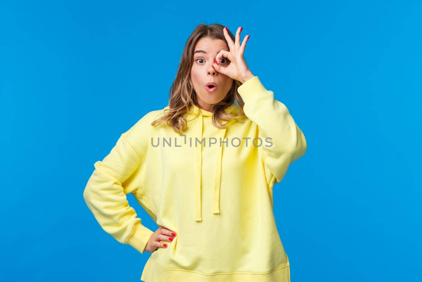 Very good, awesome. Impressed happy blond girl found something she was looking for, show okay gesture over eye and look through fingers, say wow astonished, stand blue background.