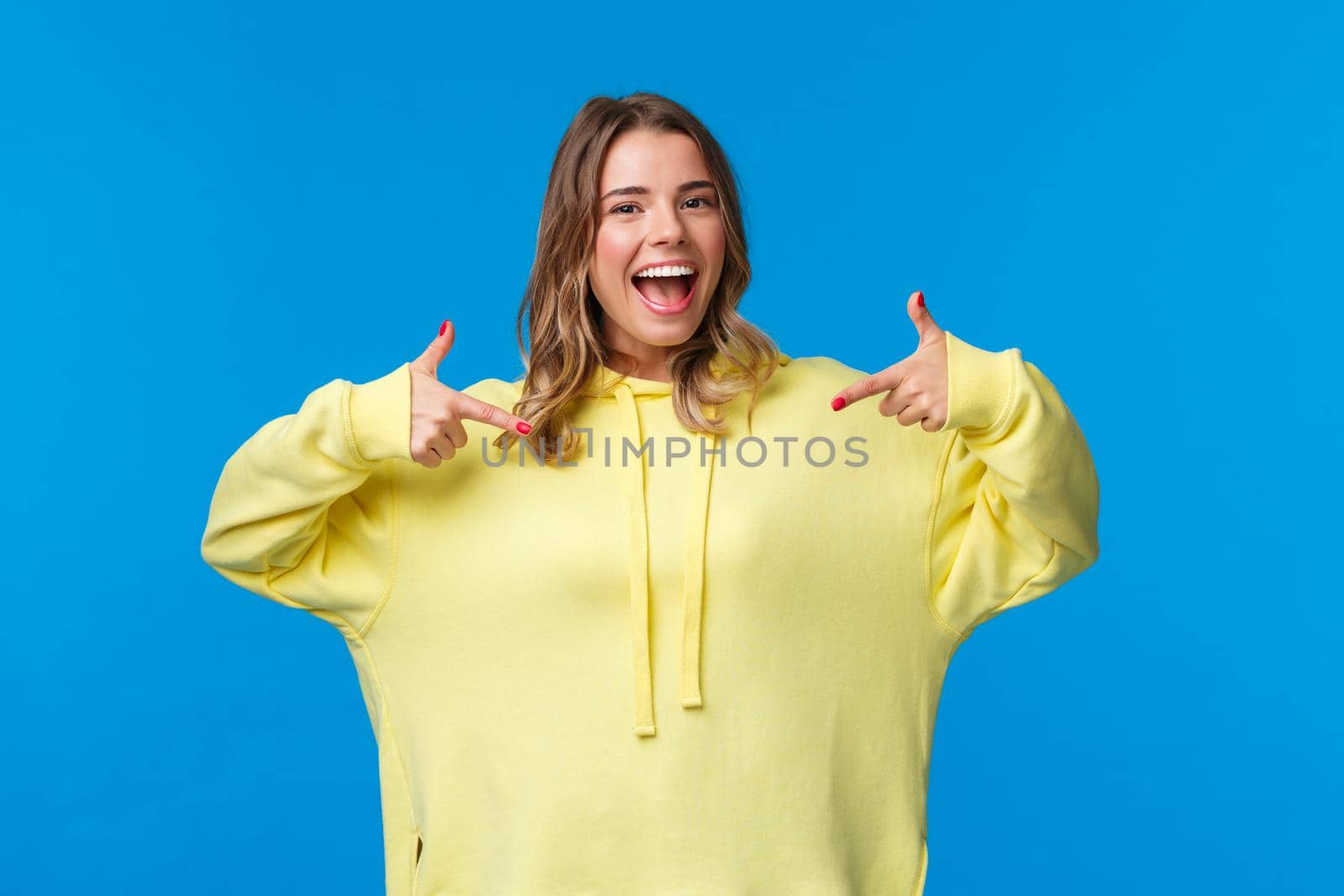 Confident, friendly-looking smiling blond girl in yellow hoodie pointing at herself, suggest own help, want volunteer or particiapte, say pick me, standing blue background.