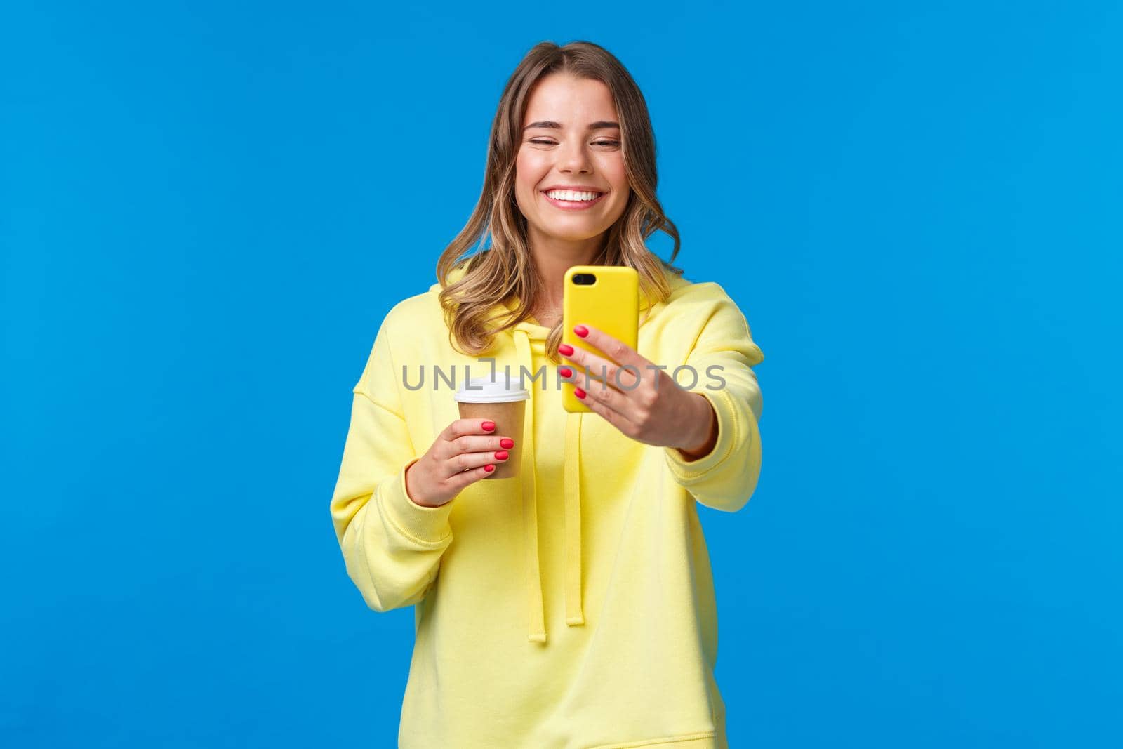 Joyful pretty feminine blond short-haired woman in yellow hoodie, taking selfie with take-away coffee from favorite local cafe, smiling smartphone camera standing with mobile phone by Benzoix