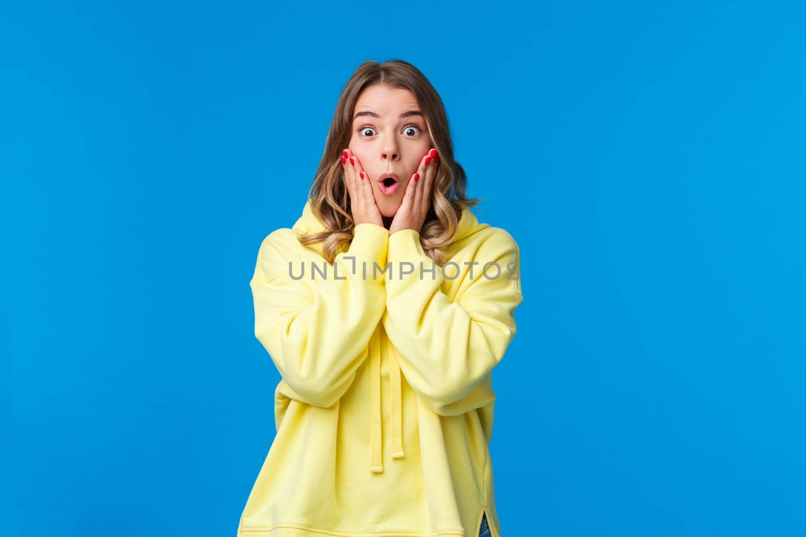 Impressed speechless young blond woman open mouth stare startled camera and say wow, touch cheeks from amazement stare disbelief and amazement, hear fantastic news, blue background.