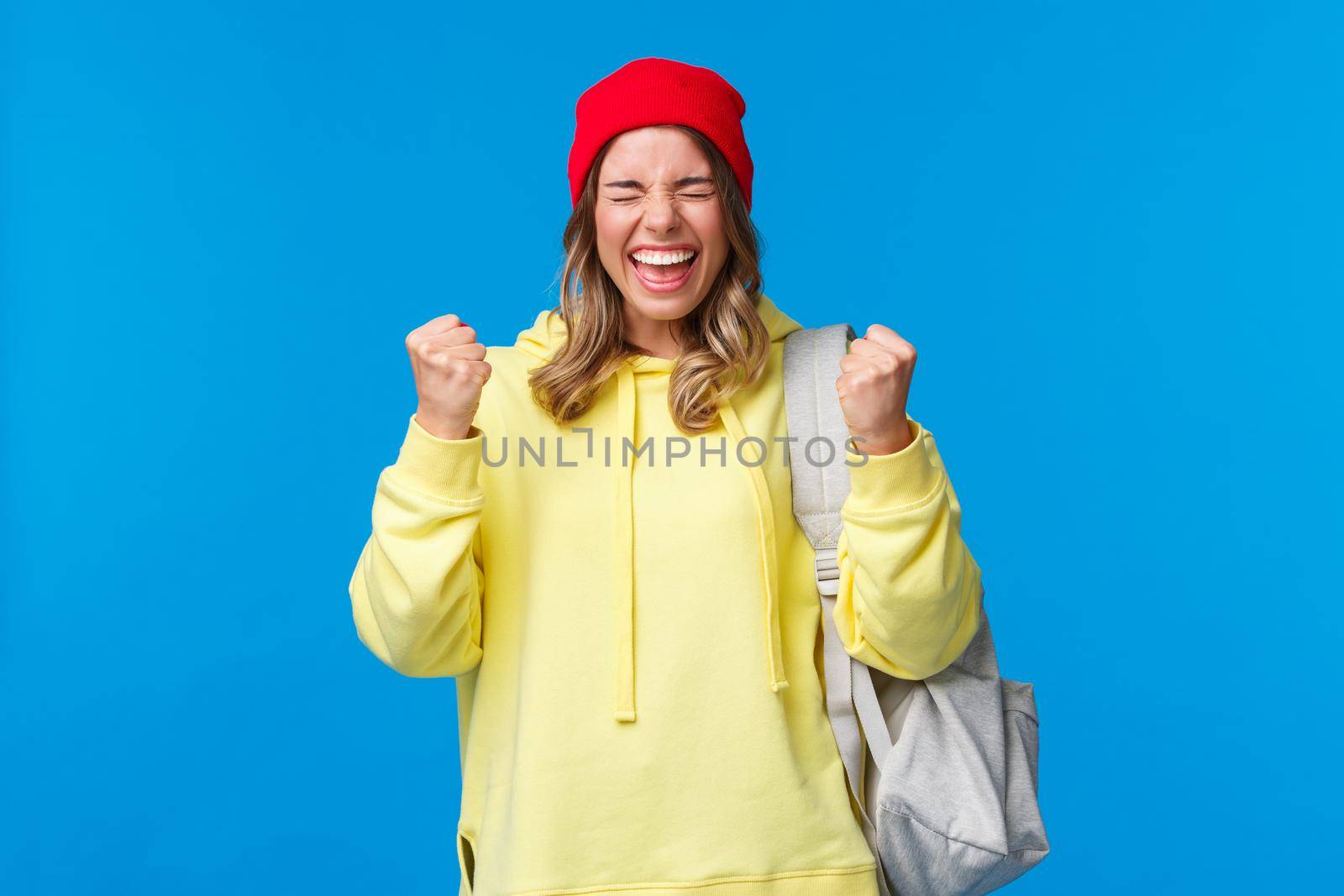 Happy cheerful blond girl in red beanie and yellow hoodie, close eyes relieved and smiling triumphing, celebrating great news, passed exams in university, fist pump like champion, winning prize.