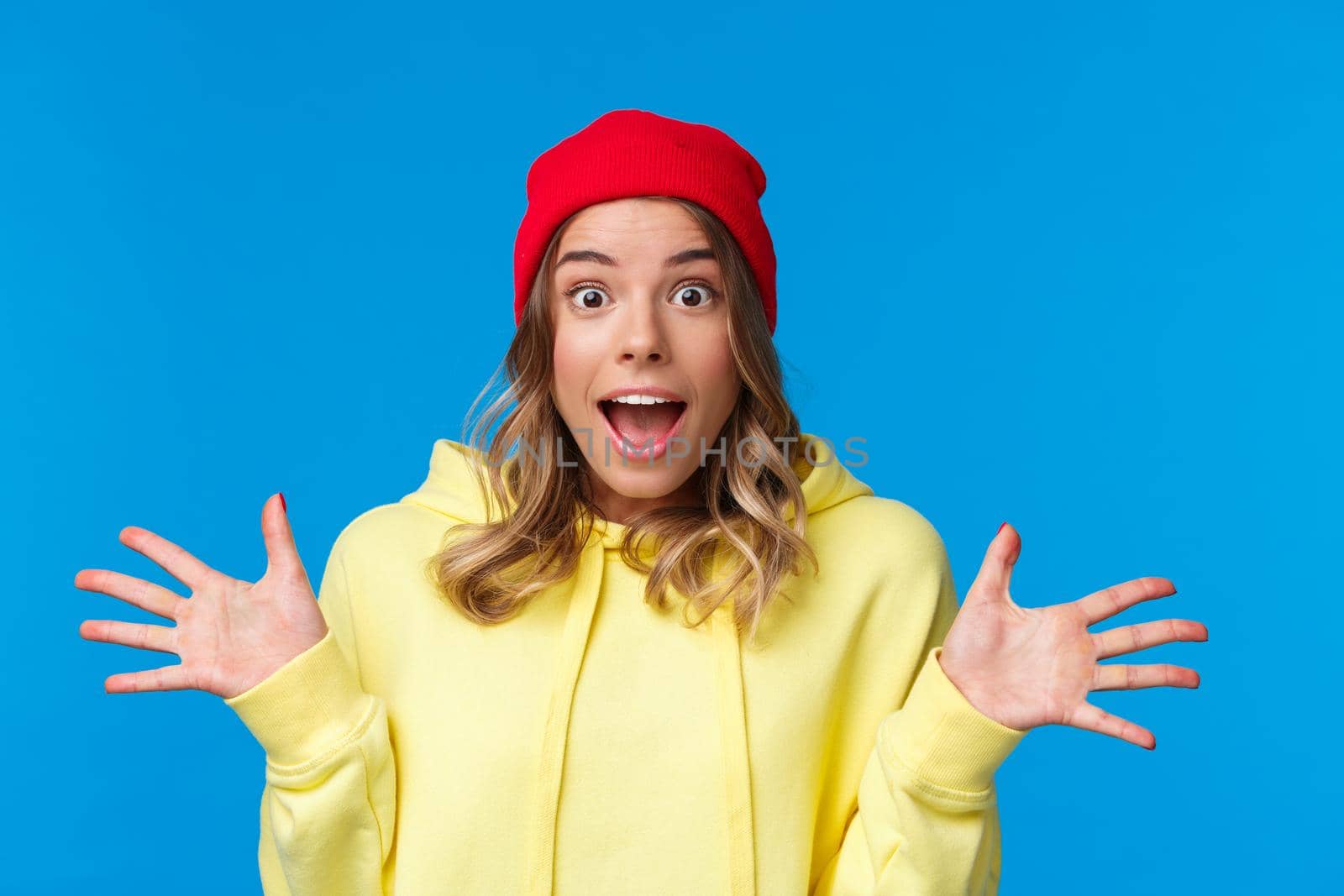 Close-up portrait thrilled and joyful amused european female in red beanie and yellow hoodie, listening to wonderful awesome news, spread hands sideways excited, stand blue background by Benzoix