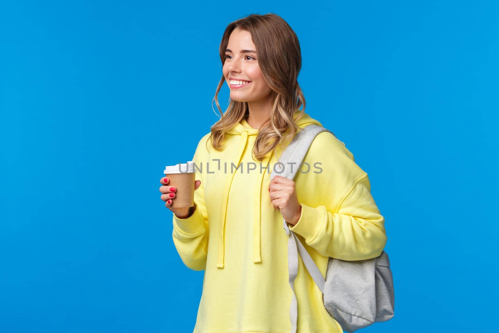 Carefree pretty young female student drinking coffee from paper cup while heading university, holding backpack and looking aside with pleased relaxed smile, lifestyle and people concept.