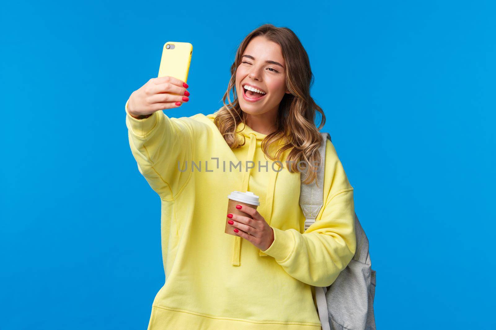 Lifestyle, university and education concept. Cheerful good-looking blond female student or traveller taking selfie with backpack and take-away coffee in paper cup, wink to mobile phone camera.