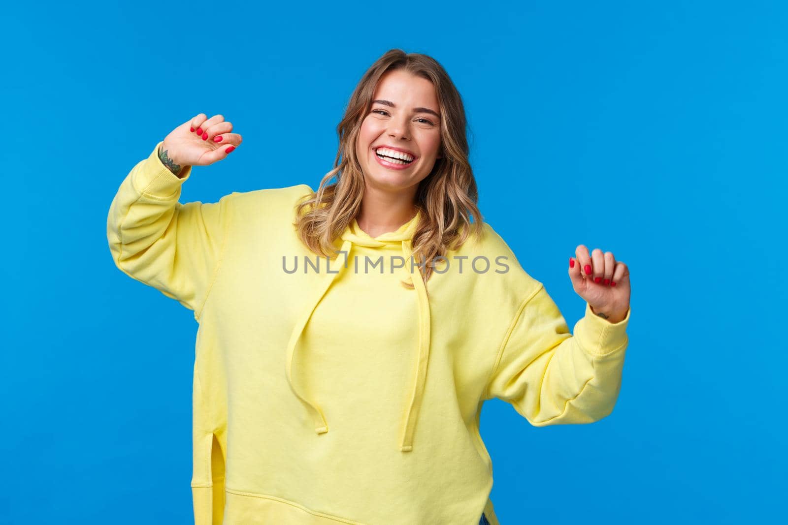 Carefree happy alluring young blond woman having fun, vibing on weekends, enjoying party as dancing and laughing over blue background, lift hands up joyful.