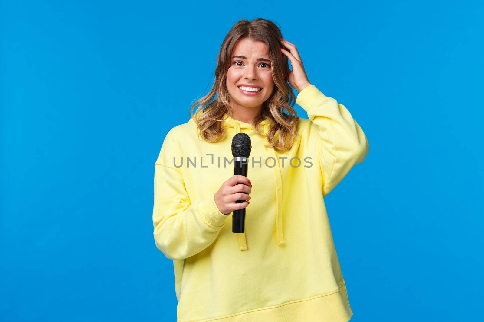 Embarrassed girl holding microphone awkward smiling and scratch head as singing song on karaoke with friends, shy perform in front of audience, standing blue background.