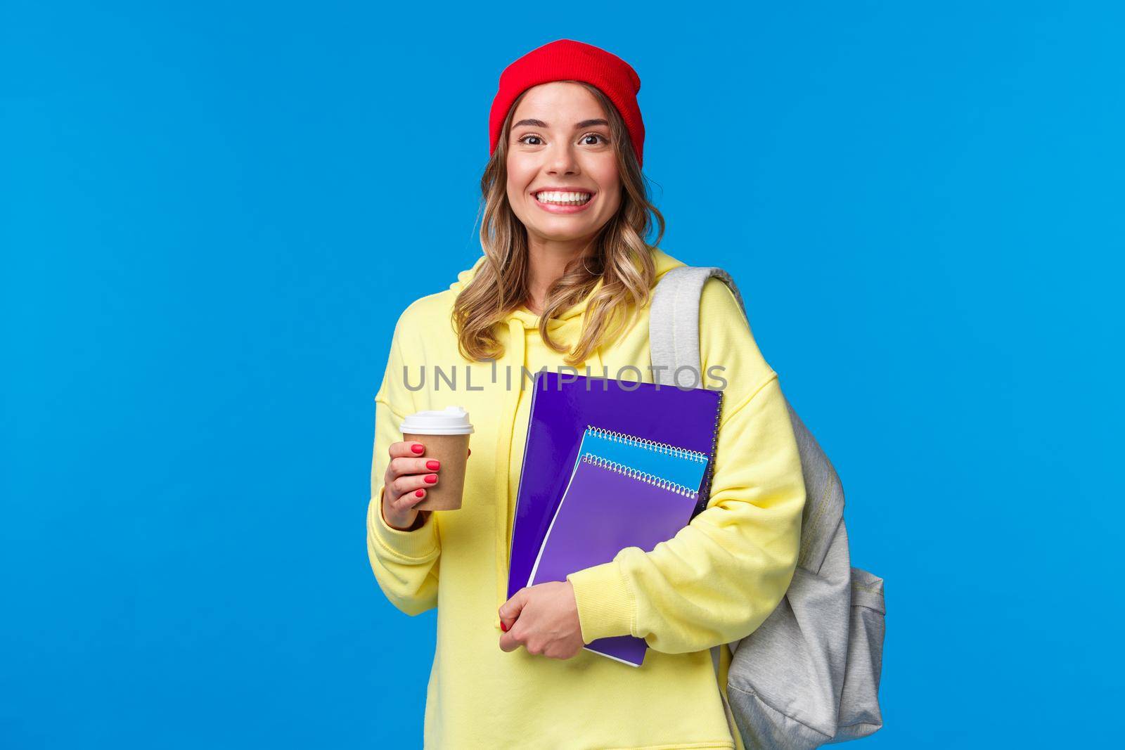 Happy excited cute girl starting senior year in college, holding notebooks and take-away coffee, carry backpack, smiling upbeat camera, wearing hipster beanie and yellow hoodie.