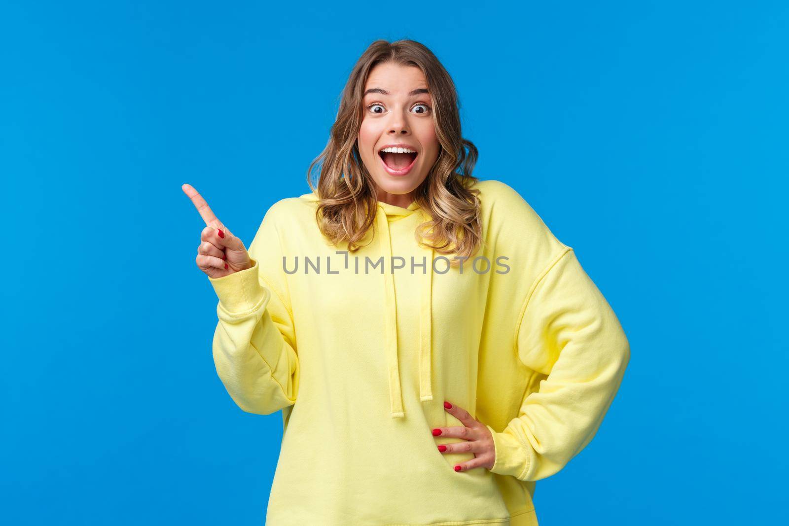 Surprised and happy, excited blond girl hear excellent news, amazing surprise, pointing finger left and looking camera with disbelief and upbeat feelings, standing blue background.