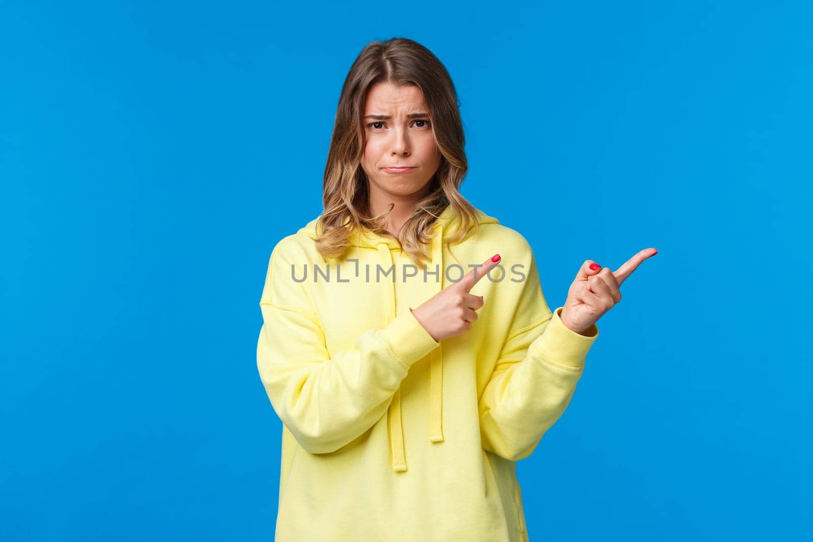 Silly insecure and sad cute caucasian female student with blond hair, sulking and smirk upset telling bad news, pointing fingers right at something disturbing and bad, blue background.