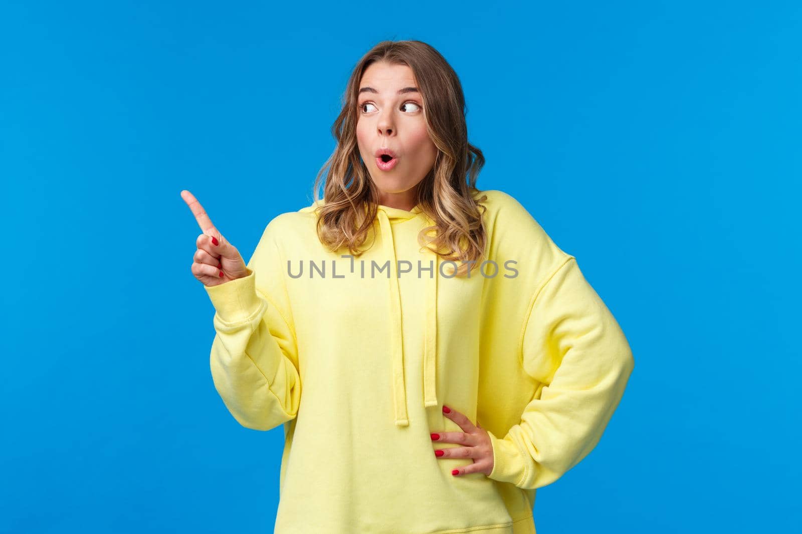 Thrilled speechless good-looking blonde female in yellow hoodie react to something shocking and interesting, pointing finger and looking left saying wow, gossiping, stand blue background.