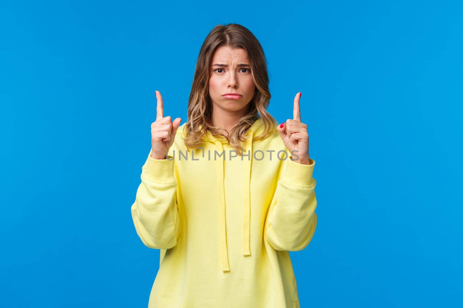Offended and gloomy, uneasy cute sulky blond girl telling about something bad happened, worst day ever, looking camera pouting and frowning insulted, pointing fingers up, blue background.