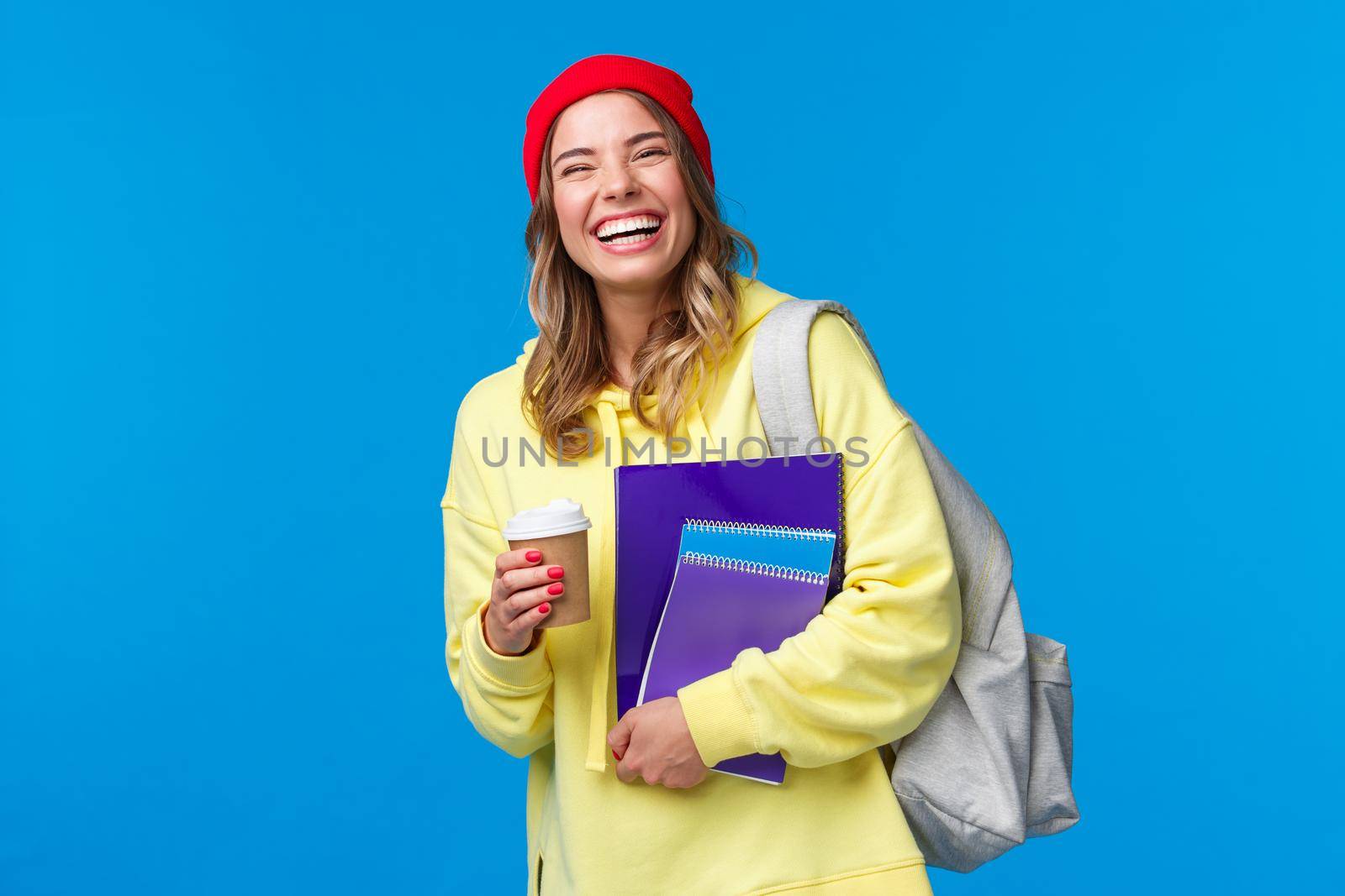 Cheerful happy female student having fun, enjoying university lifestyle, carry backpack and notebooks, drinking take-away coffee, talking to classmates, standing blue background joyful by Benzoix