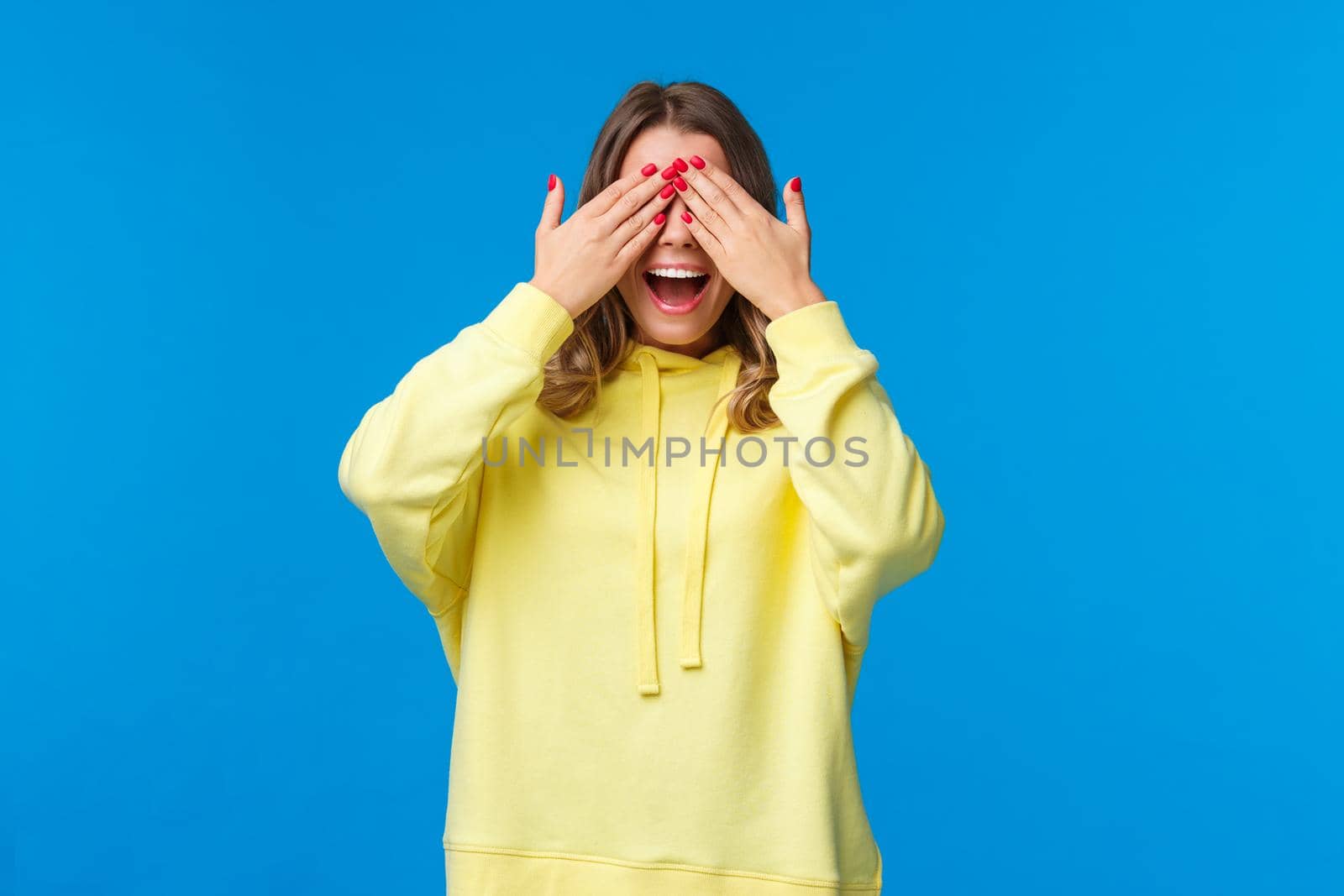 Girl waiting for friends tell her open eyes as shut it with hands and count ten during b-day party, girlfriend bring surprise gift, smiling with excitement, playing peekaboo, stand blue background.