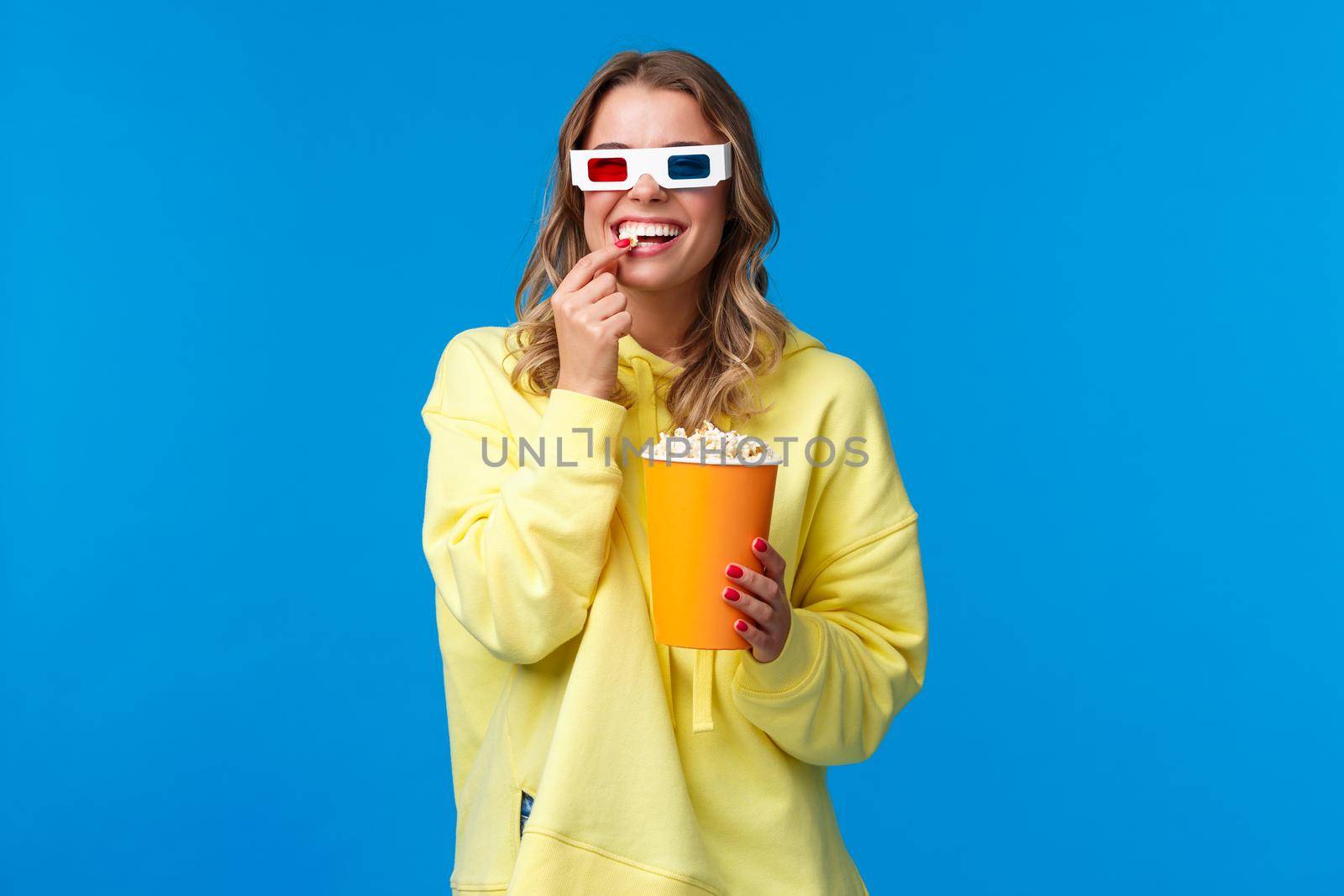 Leisure, fun and youth concept. Happy entertained smiling blond girl in 3d glasses, eating popcorn and laughing as watching funny comedy, go cinema watch movie with friends, blue backgorund.