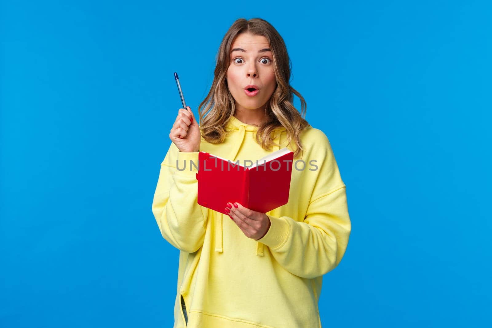 Girl doing homework finally have great plan, made-up idea for story as writing it in red notebook, raise pen eureka gesture, looking amazed camera, standing blue background in yellow hoodie.