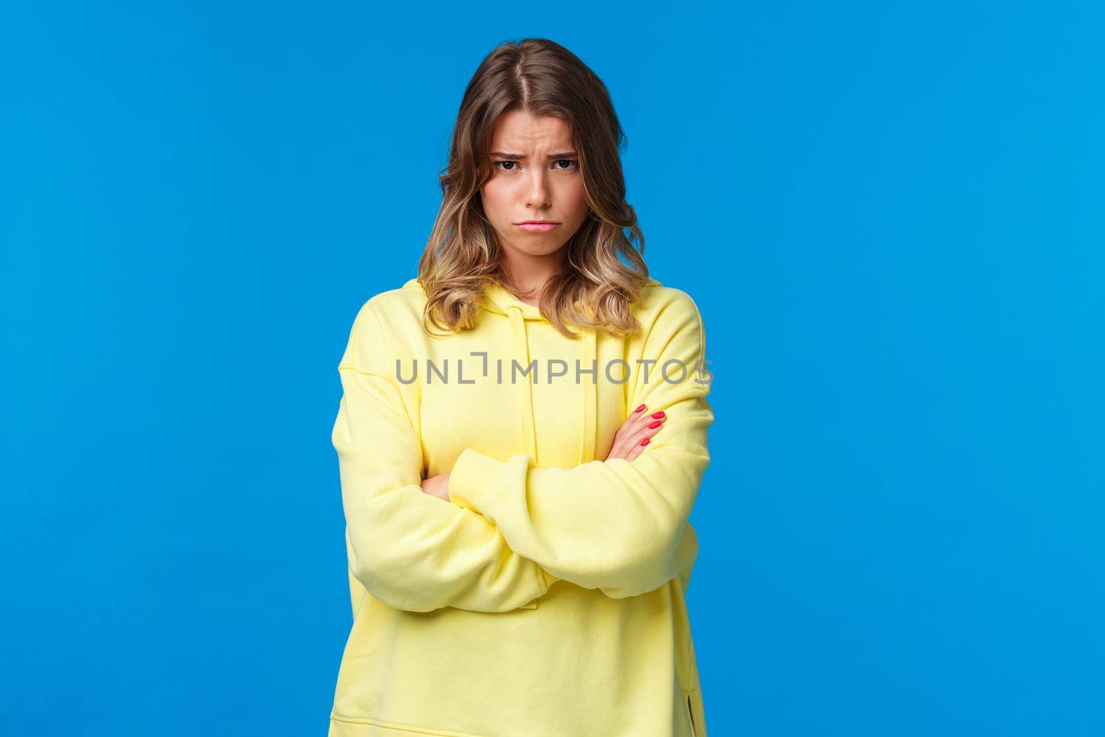 Gloomy and moody cute blond teenage girl look from under forehead and sulking at camera, feeling offended or upset, cross hands chest insulted waiting apology, blue background by Benzoix