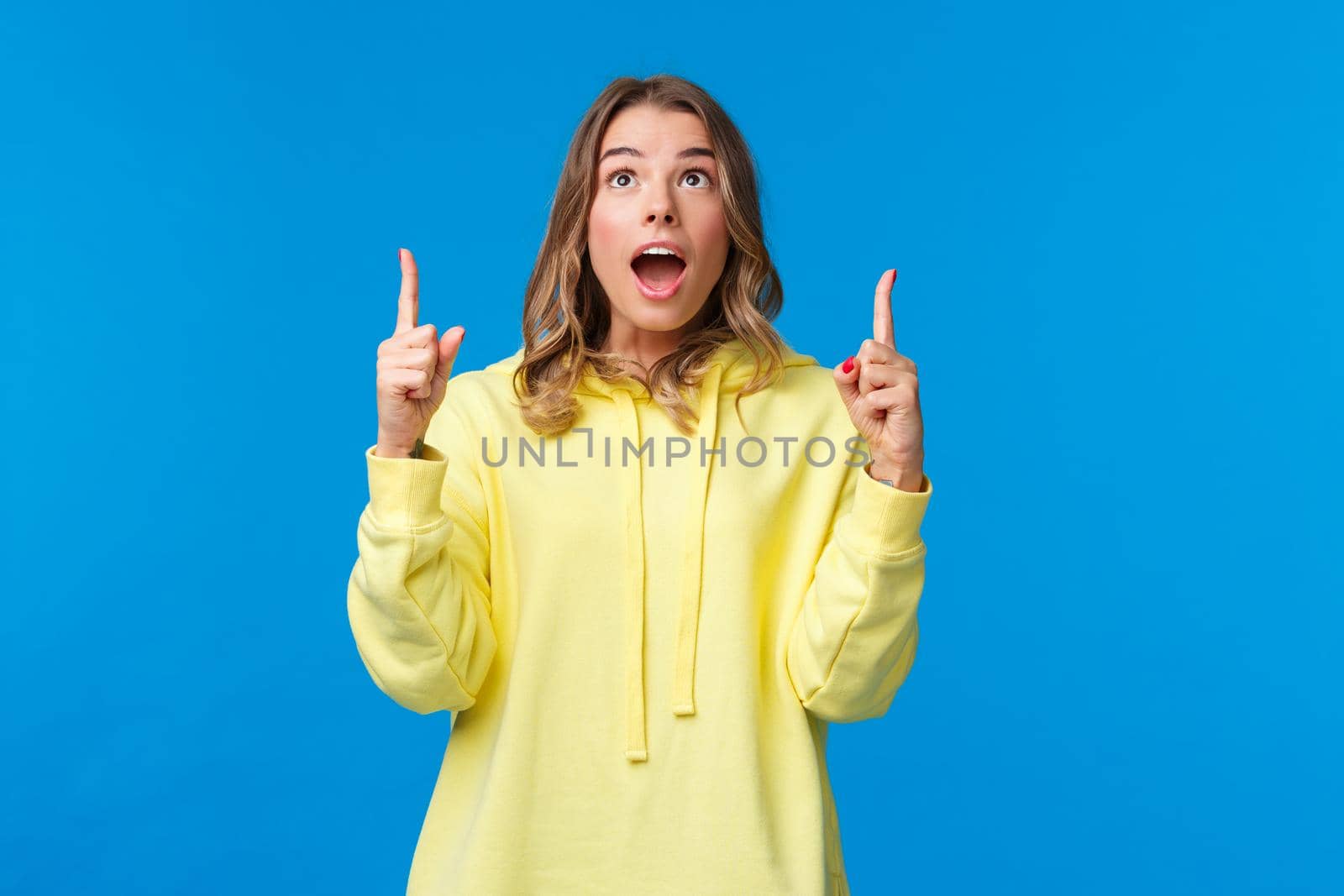 Fascinated and amused young blond girl drop jaw and staring speechless, pointing fingers up at something incredible and awesome, watching amazing performance, stand blue background by Benzoix