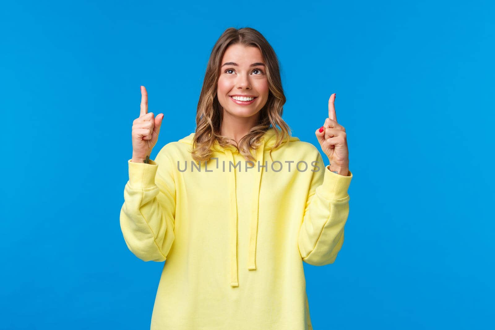 Happy alluring caucasian female with blond hair in yellow hoodie, searching career opportunities, found something interesting, looking and pointing fingers up, smiling pleased.