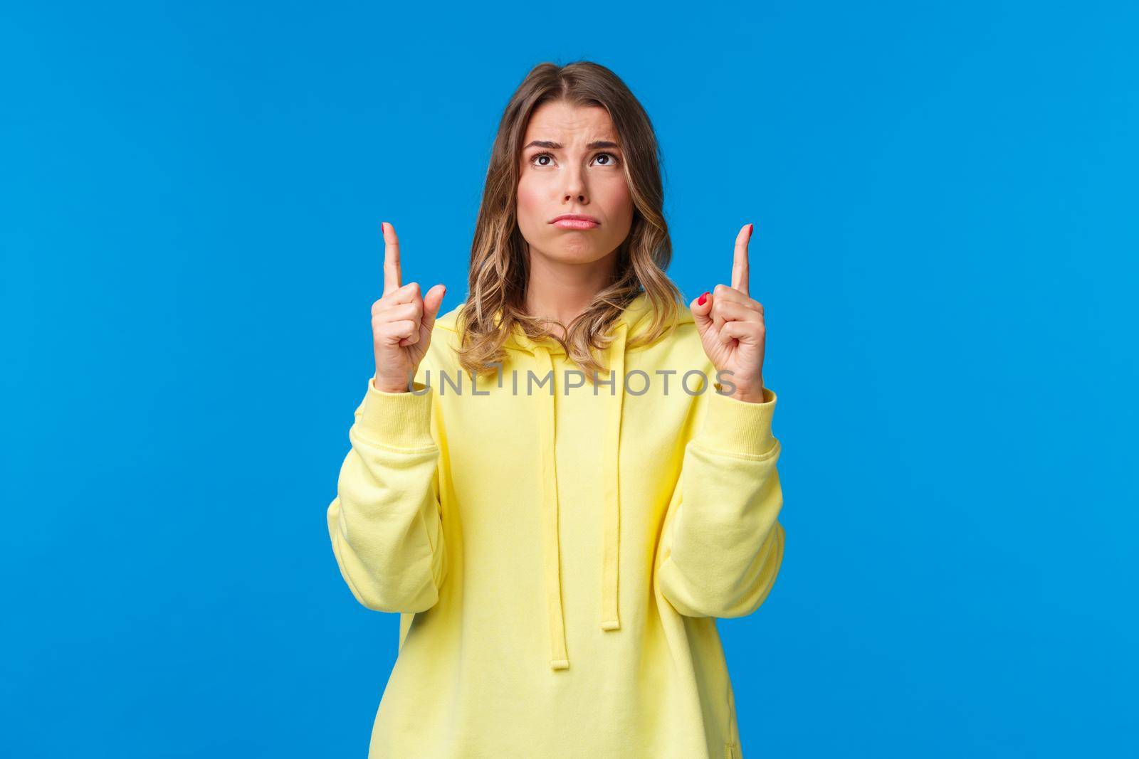 Jealous and gloomy cute blond female student feel regret not having something she desires, sobbing frowning sad as looking pointing up, standing over blue background.