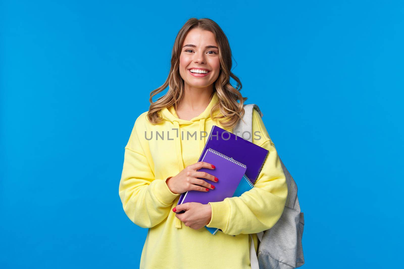 Cheerful pretty blond girl smiling at camera, carry backpack and notebooks, papers for studying, learning new language at courses, standing joyful over blue background. Education and people concept