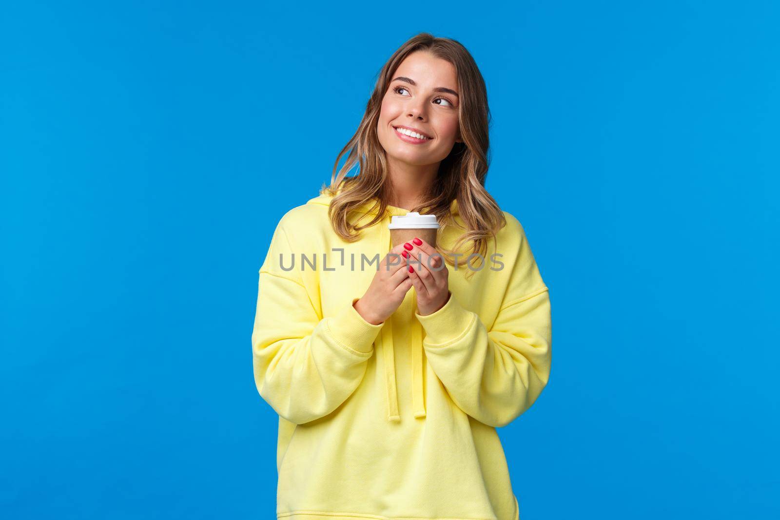 Dreamy cute blond european girl in yellow hoodie looking up romantically and thoughtful, smiling to herself as imaging something kawaii holding paper cup drinking coffee, blue background.