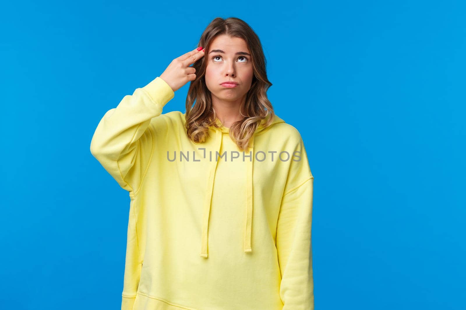 Gosh just kill me. Bored and pressured, stressed young blonde girl in yellow hoodie, make pistol gesture near head, shooting from gun and sighing as feel bothered and troubled, blue background.