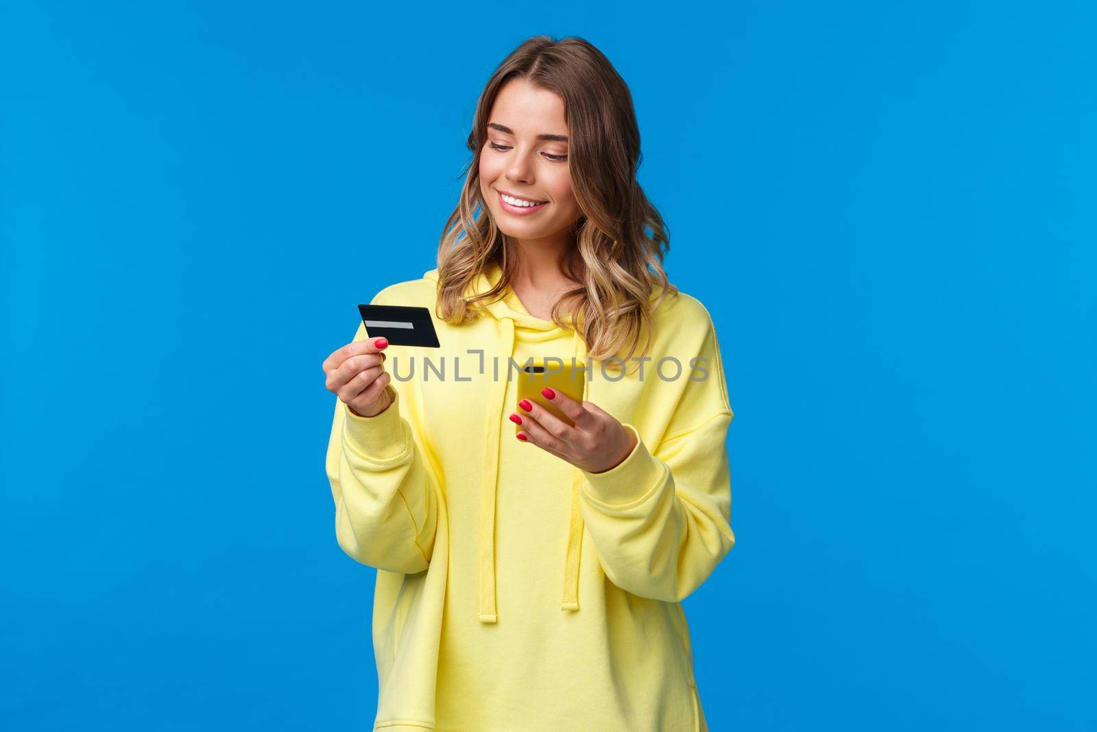 Young caucasian female with blond short hair using mobile phone and insert digit numbers of credit card to pay for her online purchase, shopping in internet store, smiling pleased.