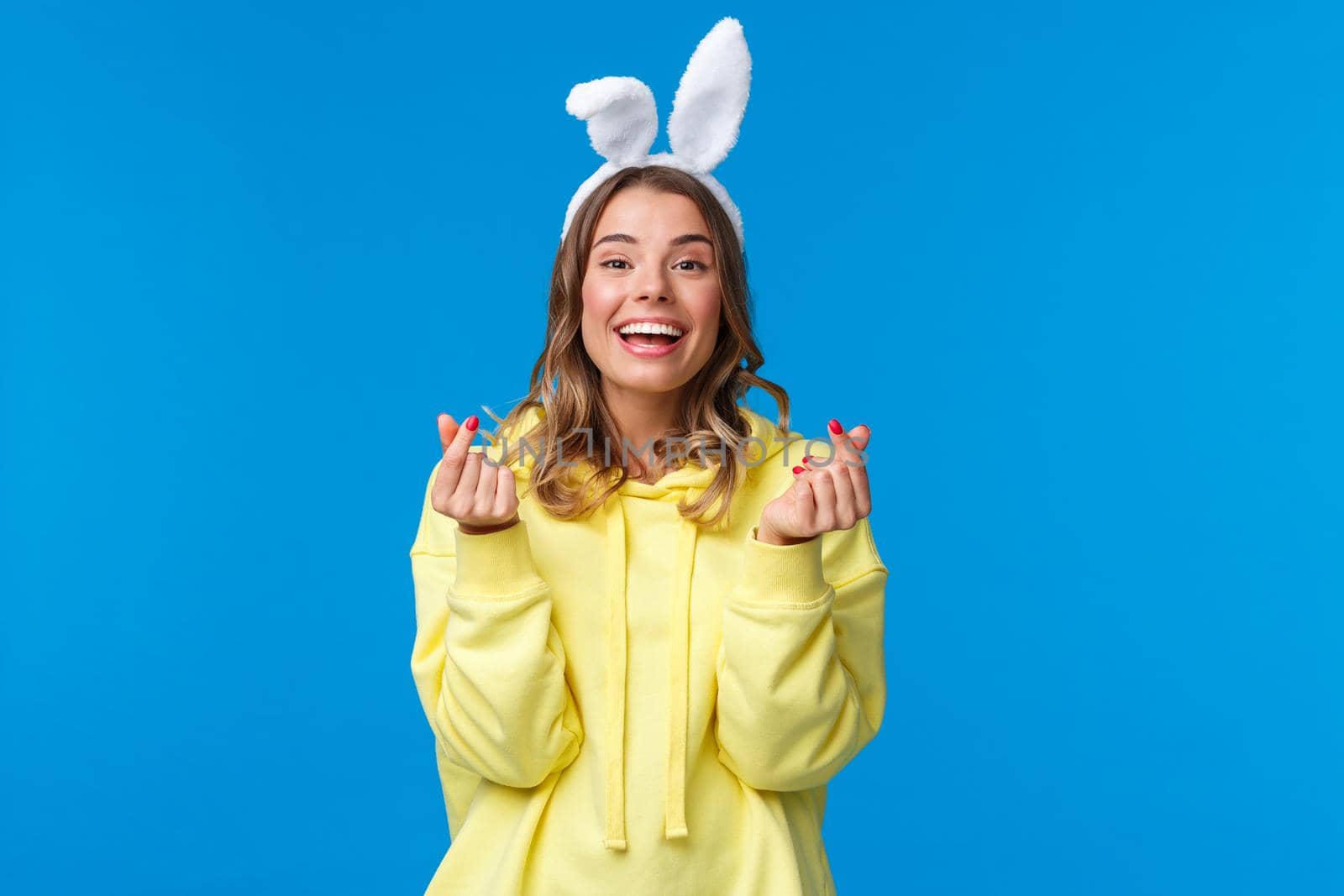 Holidays, traditions and celebration concept. Cheerful european girl celebrating Easter day, wearing rabbit ears show korean hearts sign and smiling joyfully, stand blue background.