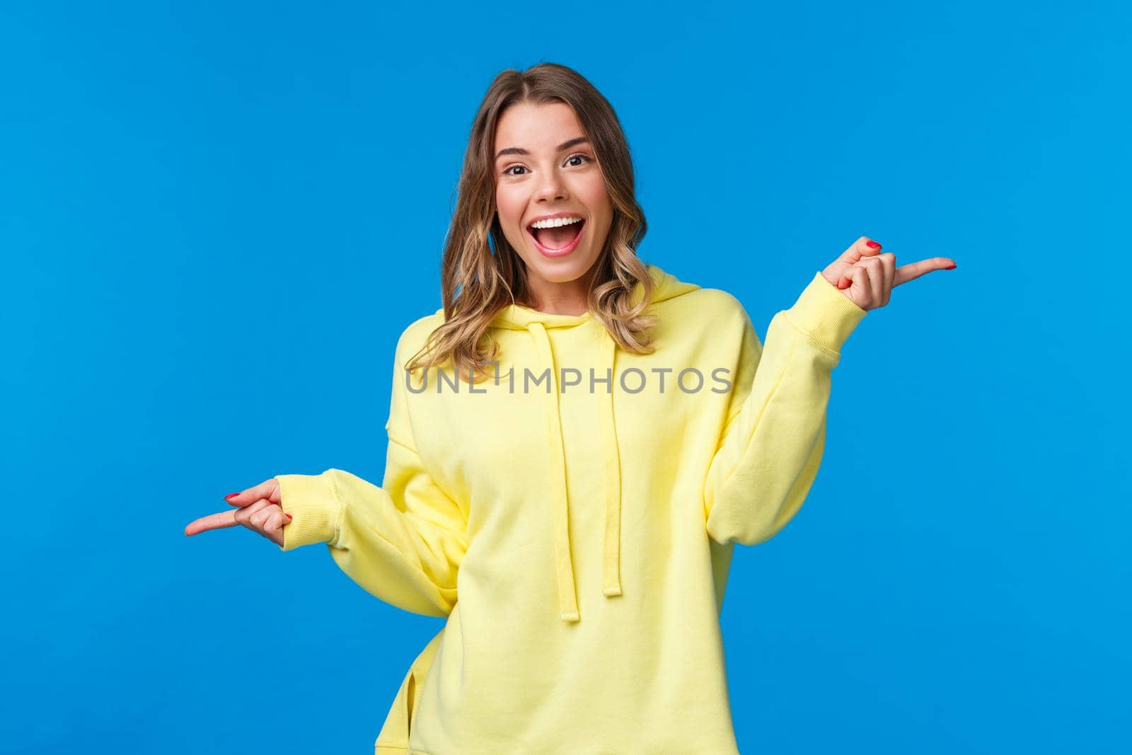 Why not try both. Carefree happy smiling young blond girl suggest two variants, pointing fingers left and right as dancing and looking camera cheerful, introduce products, blue background.