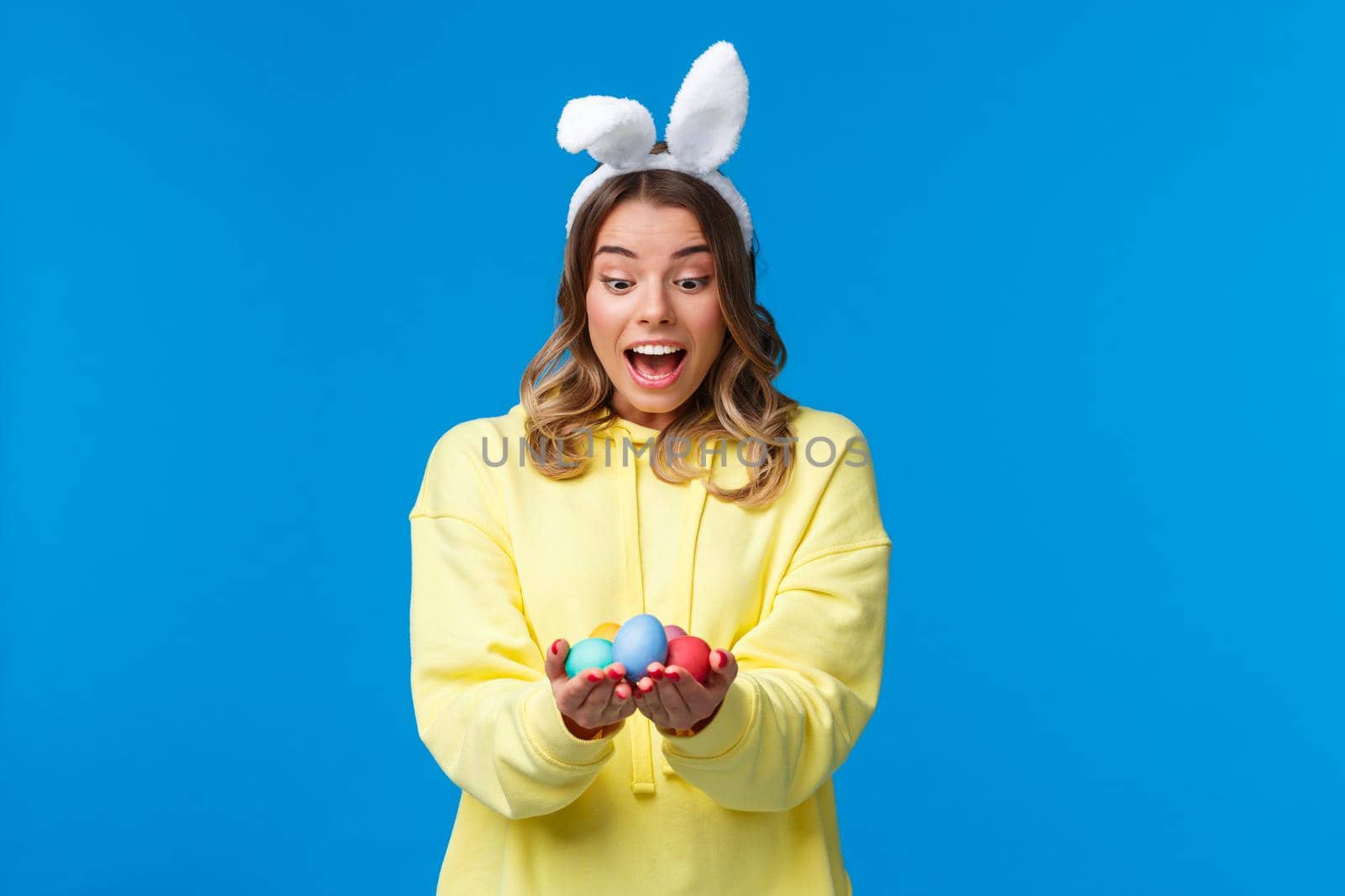 Surprised emotive cute blond caucasian girl playing holiday game, found Easter eggs look amused, wearing rabbit ears as celebrating religious holiday on sunday, blue background by Benzoix