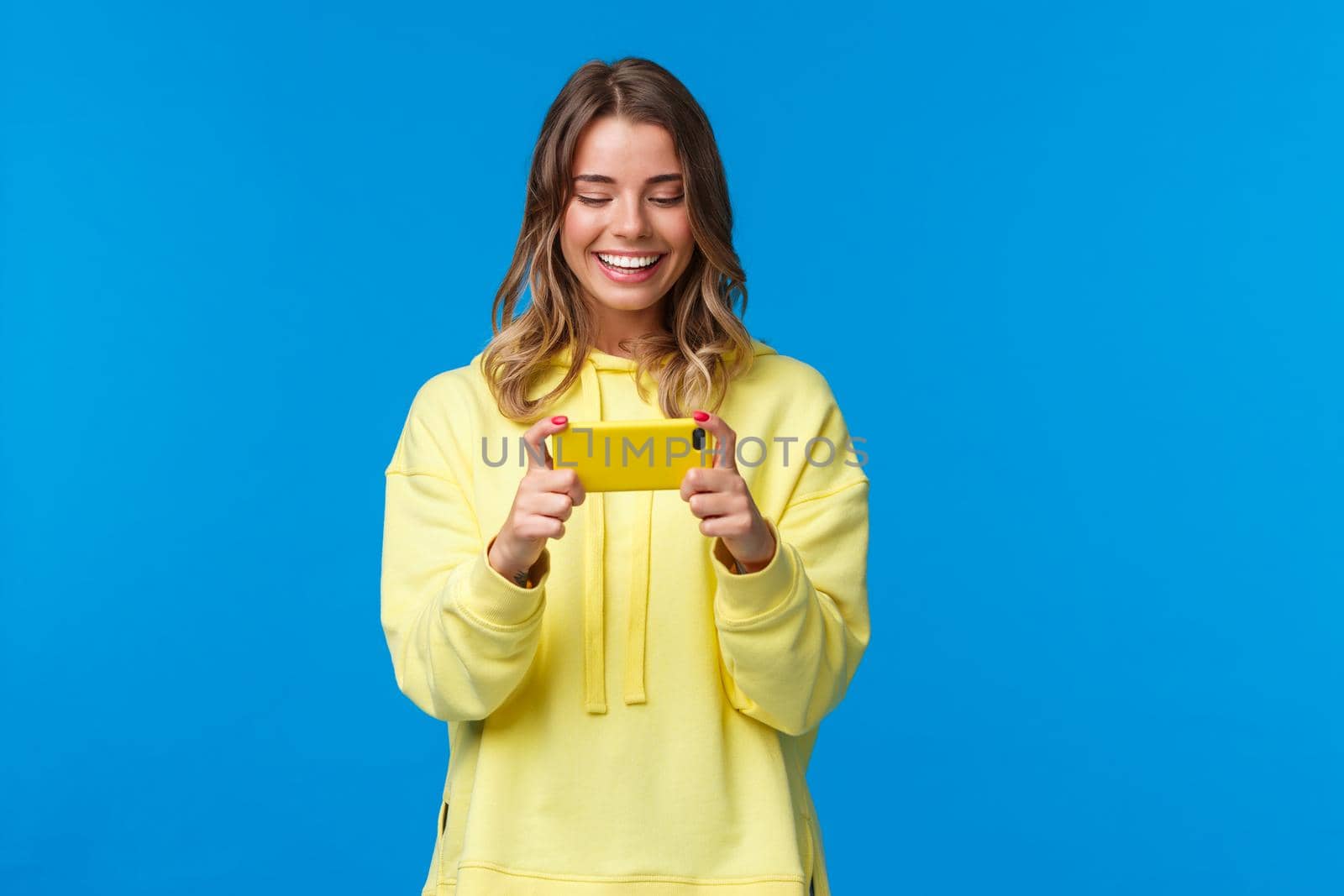 Time to relax on lunch break. Carefree good-looking hipster girl in yellow hoodie using mobile phone to play, holding smartphone horizontal position and smiling as passing easy game level.